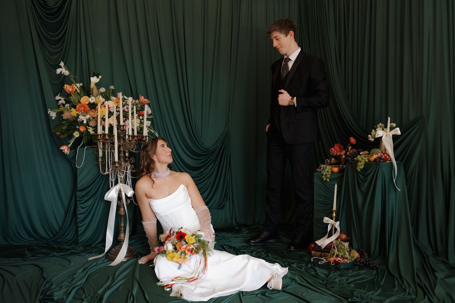 Bride in white gown sits on green draped floor holding bouquet, groom in black suit stands beside floral arrangements.