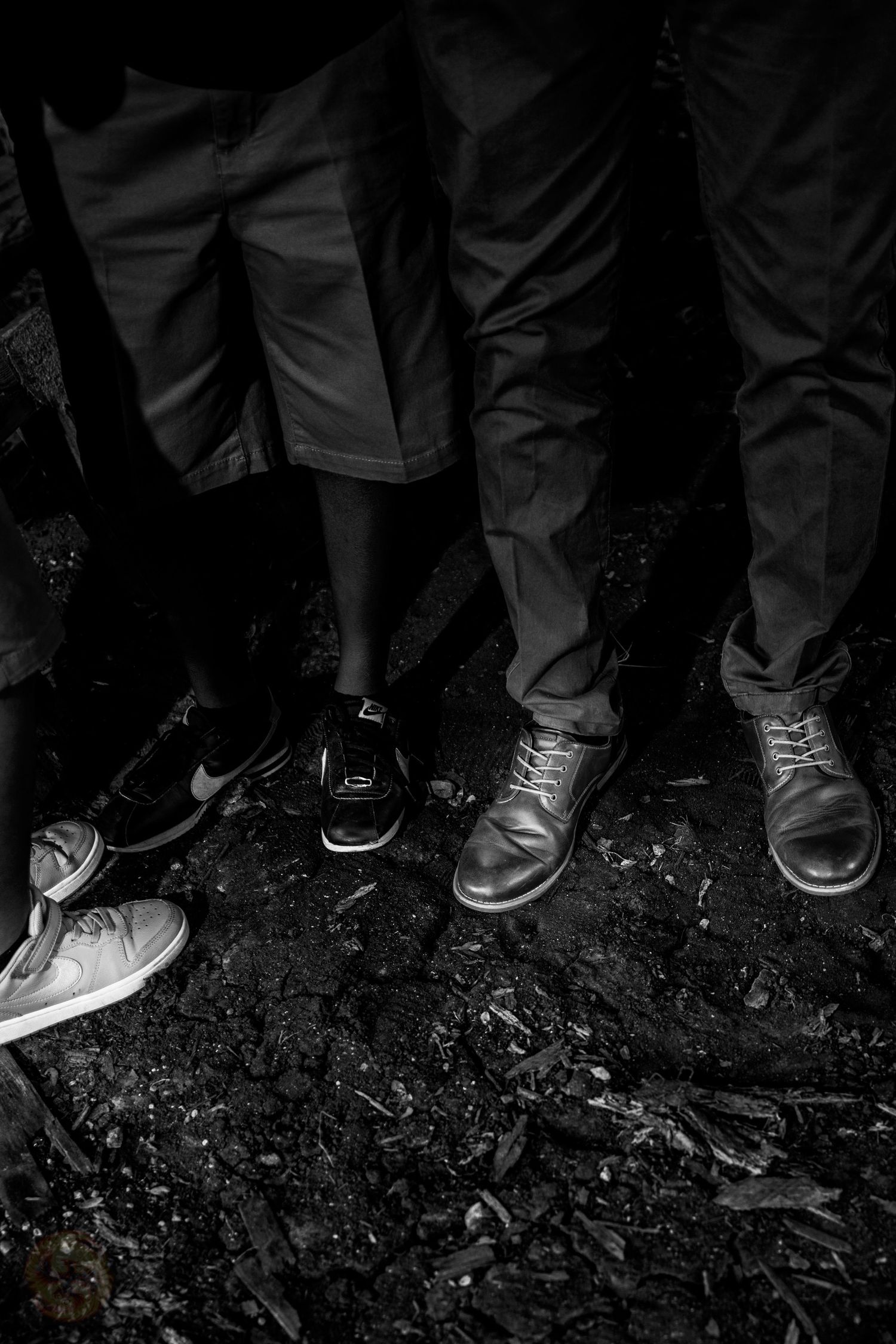 Black and white photo of people's shoes and feet lined up on dirt ground.