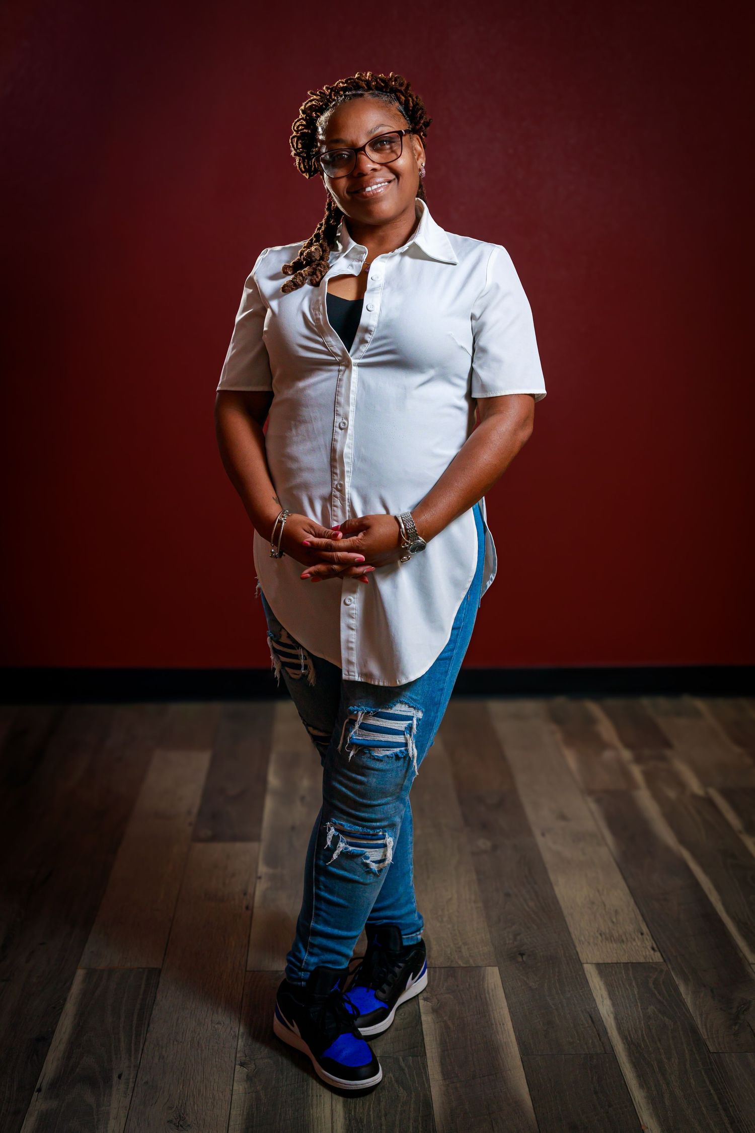A person wearing a white button-down shirt and ripped jeans stands confidently on hardwood floors against a burgundy wall.