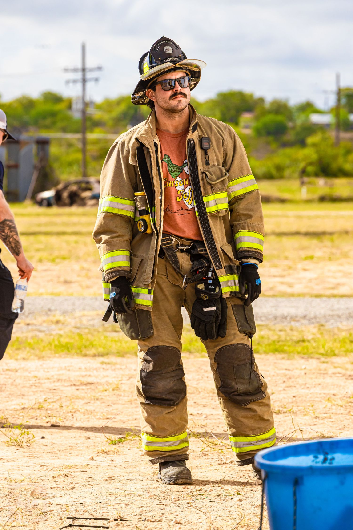 Firefighters in full gear walking across dusty training ground with equipment.