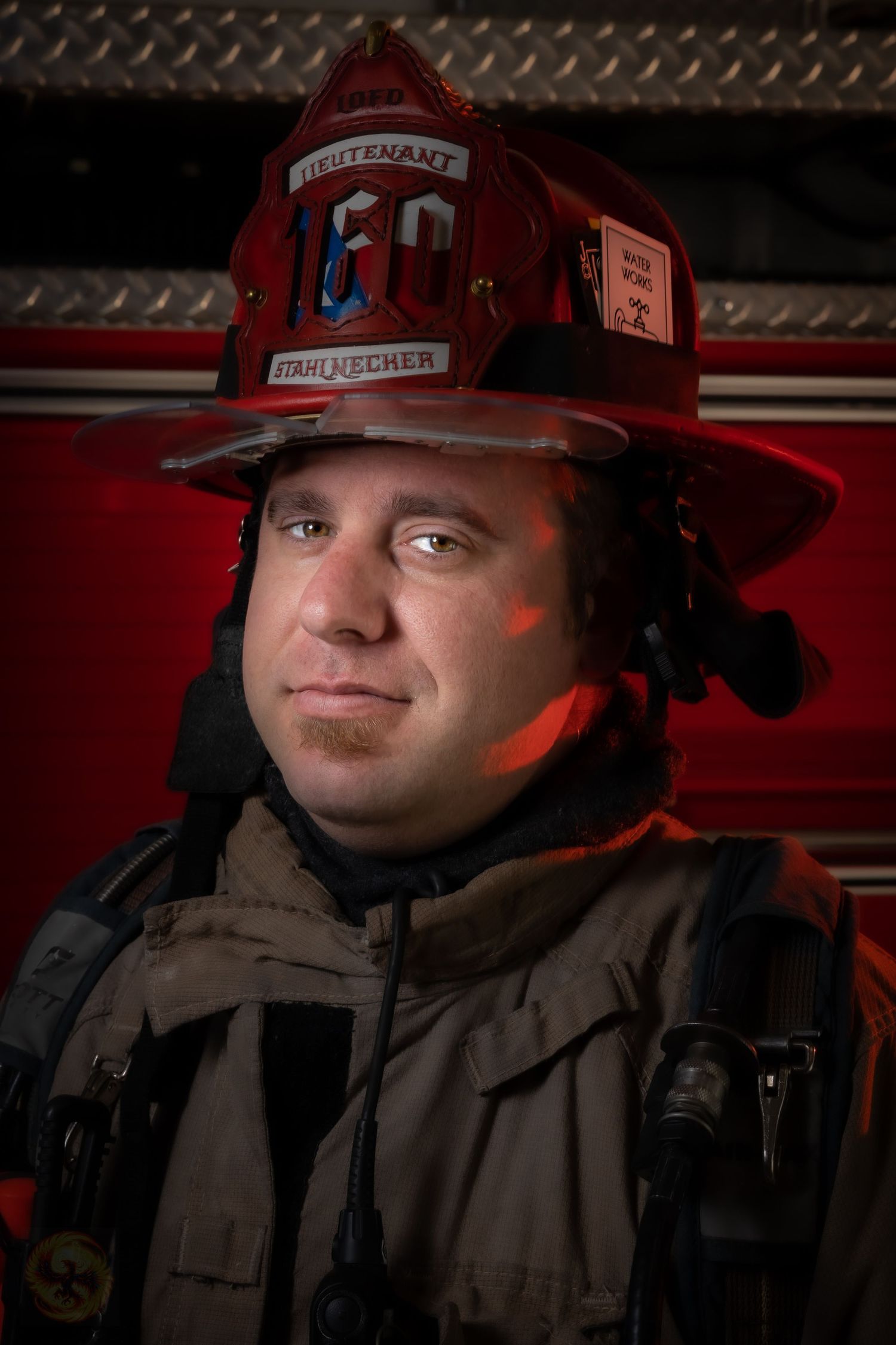 Firefighter in uniform and helmet poses against dark background with red lighting.