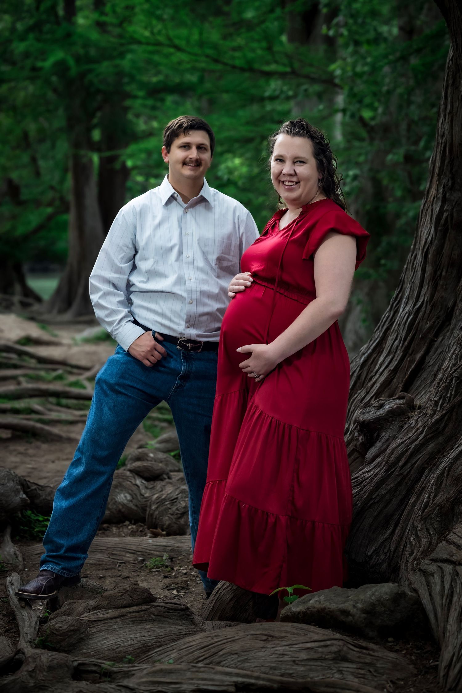 A couple poses together in a wooded forest setting wearing red dress and white shirt with jeans.