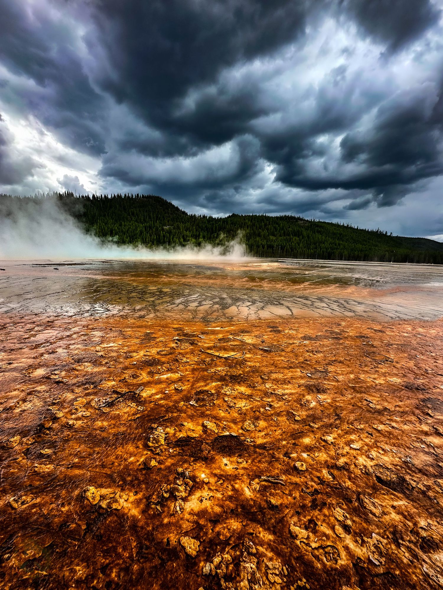 Dramatic storm clouds loom over a vibrant orange thermal pool at Yellowstone National Park.