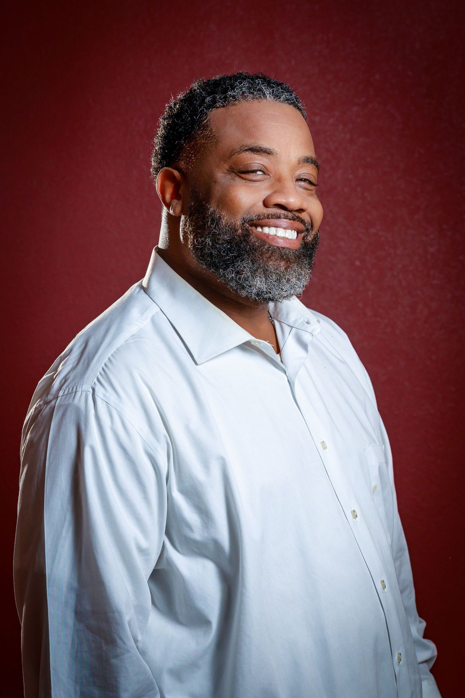 A person wearing a white button-down shirt smiles warmly against a burgundy background in a professional portrait series.