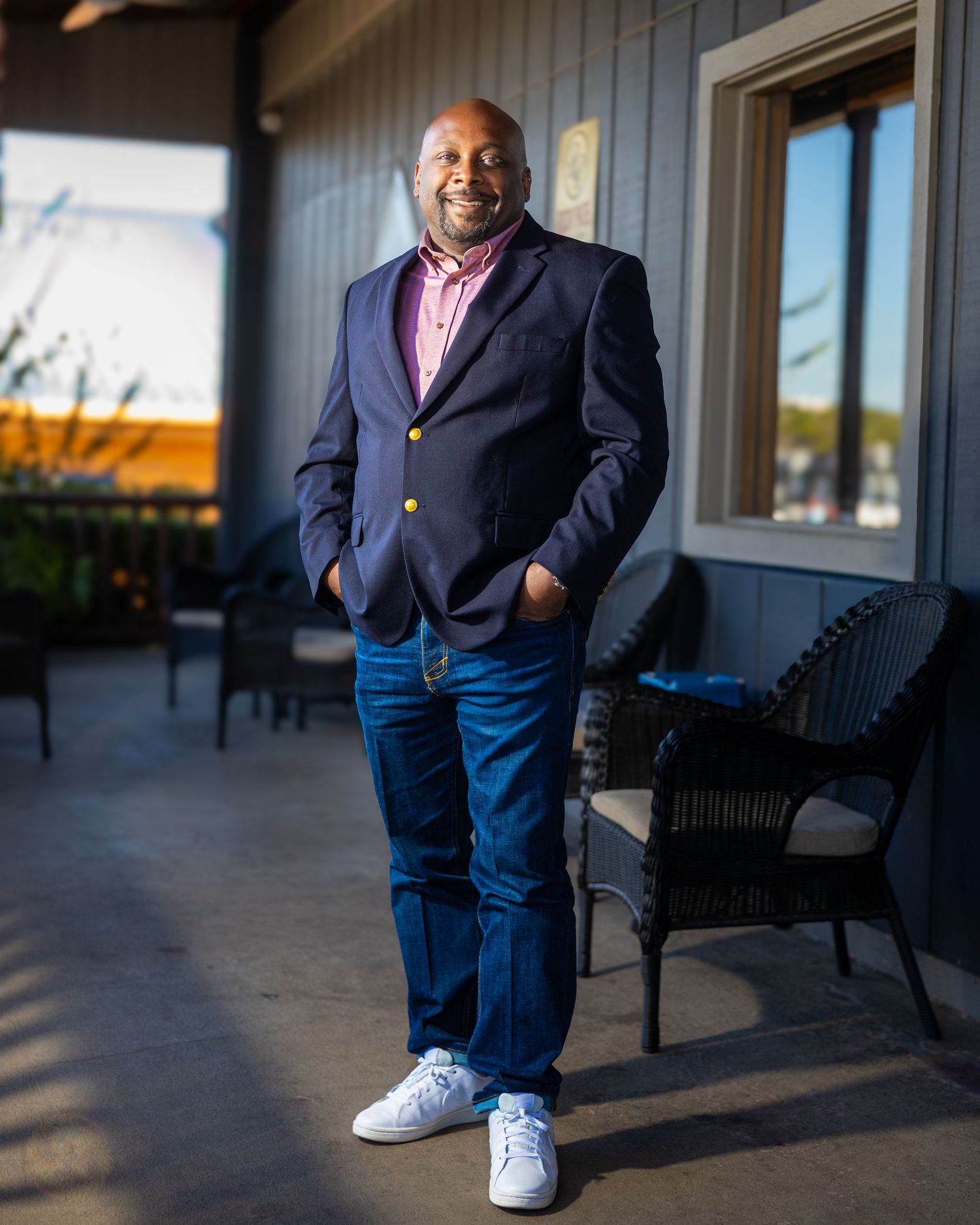 Person in navy blazer and jeans poses on outdoor porch in evening light.