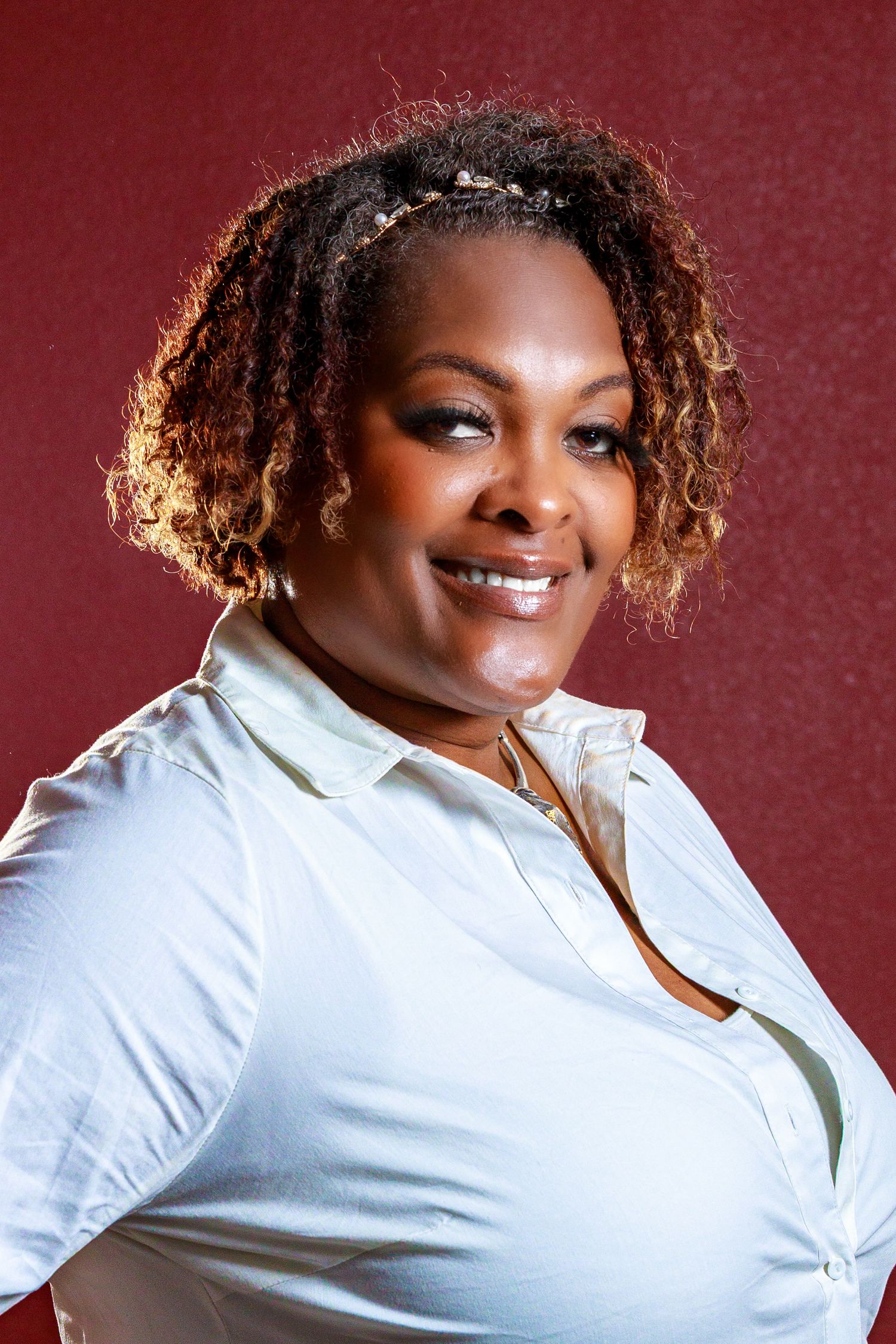 Professional portrait against burgundy background showing joyful expression and curly hair in white button-up shirt.