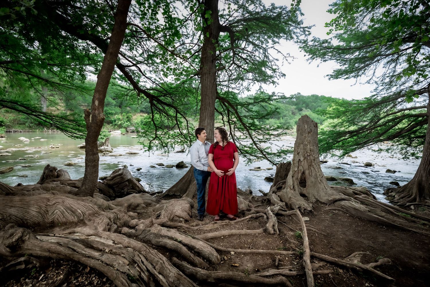 A person in a red dress poses near exposed tree roots along a rocky riverbank in a scenic forest setting.