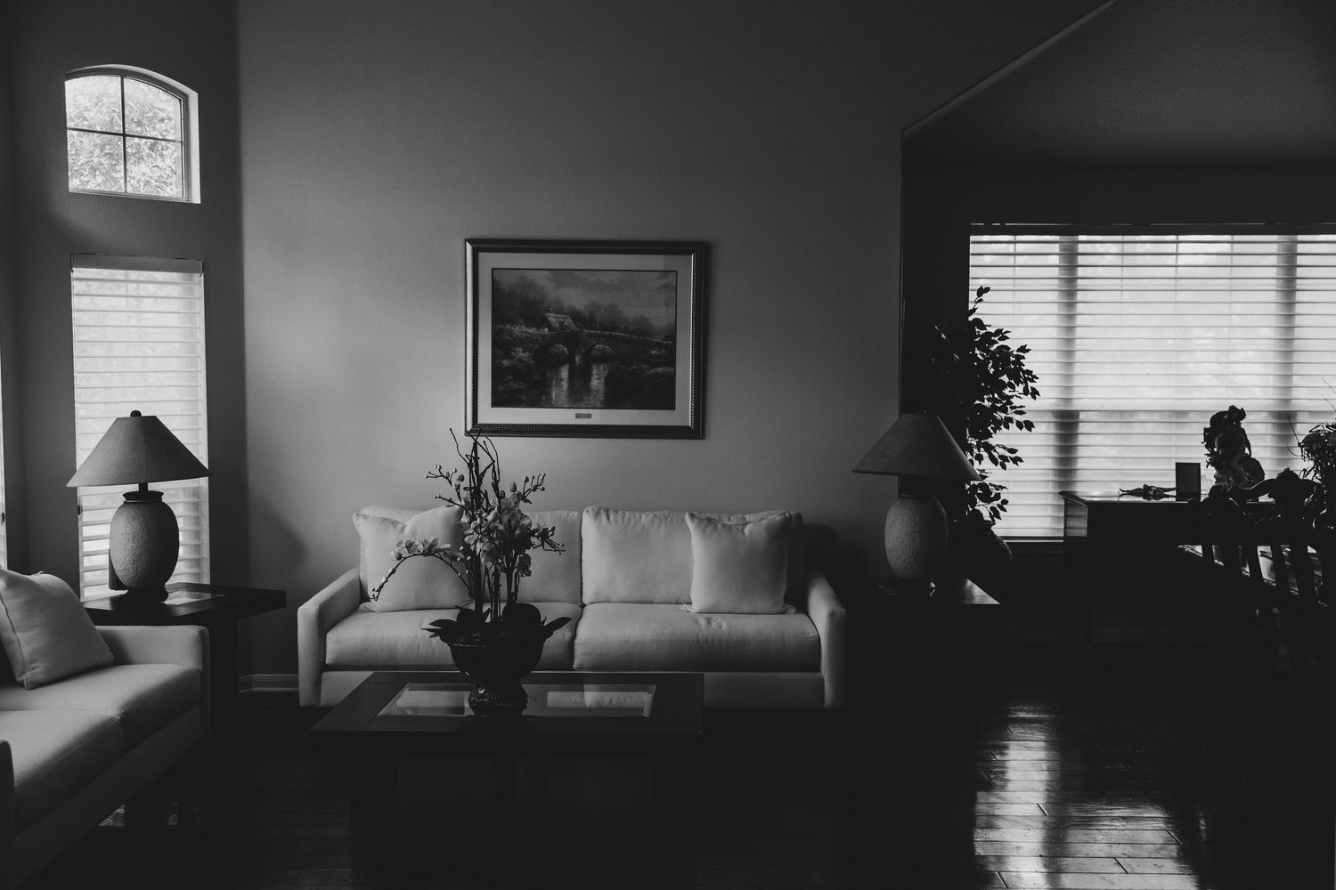 Black and white interior of a living room with a sofa, table lamp, framed artwork, and window with natural light filtering in.