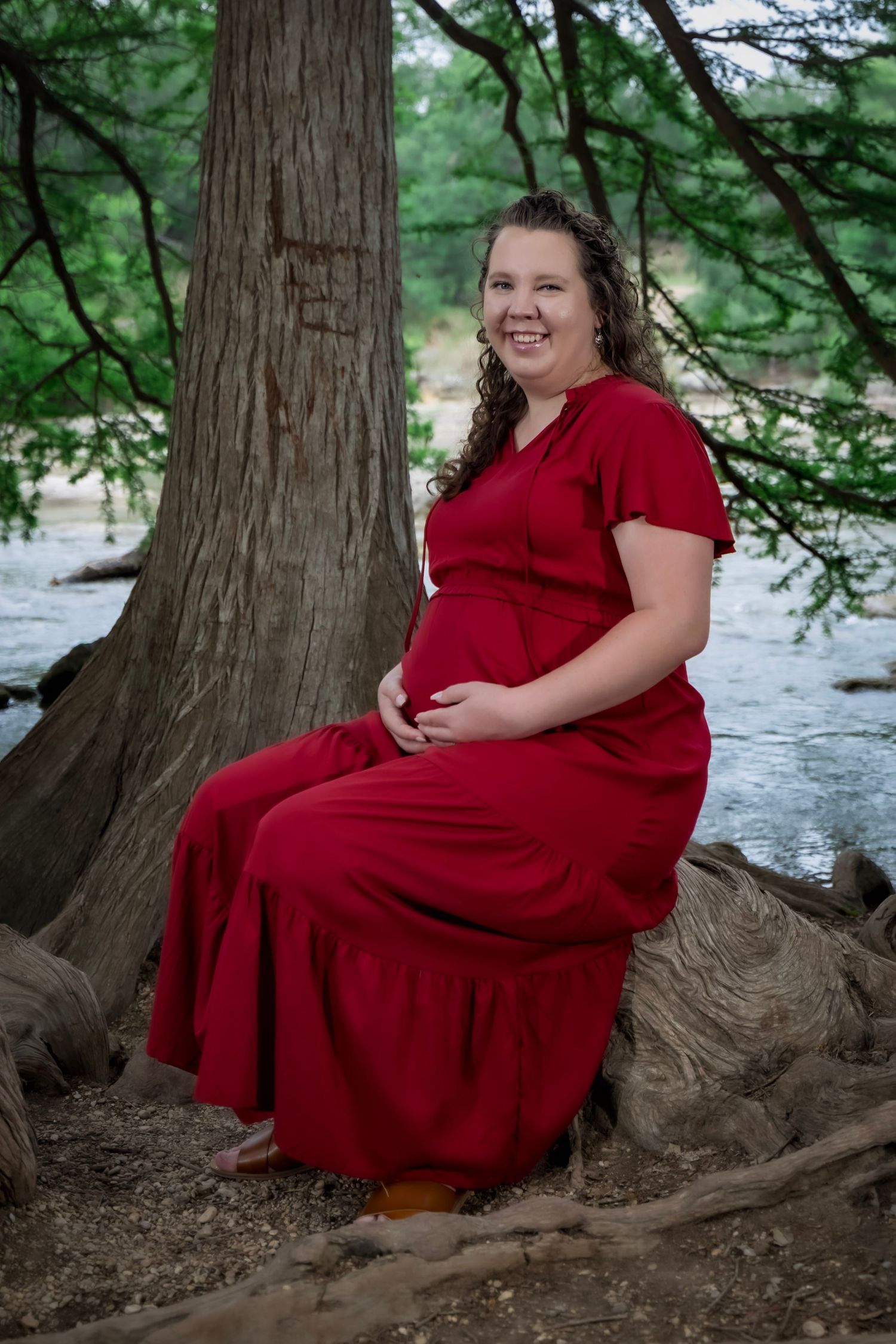 A person in a red dress poses on tree roots by a river in a scenic woodland setting.
