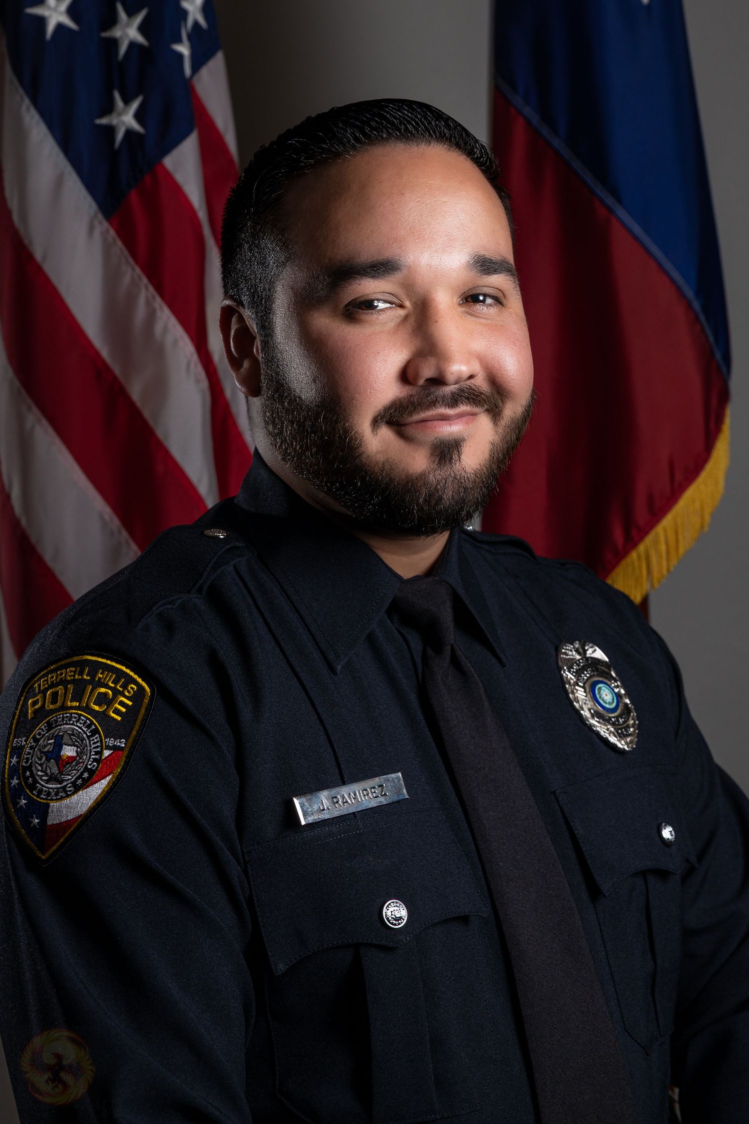 Law enforcement officer in formal uniform posing against backdrop with American and state flags.