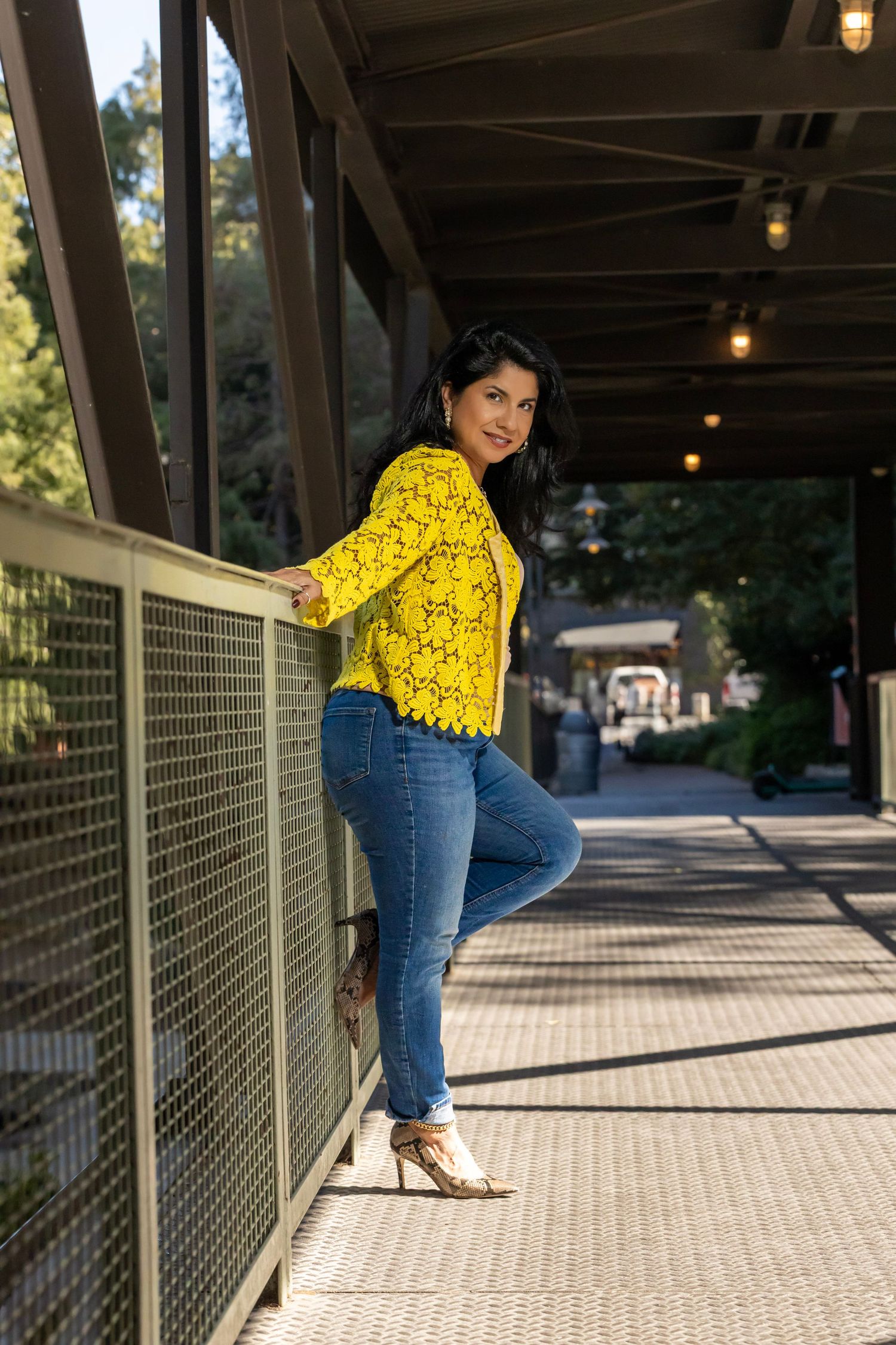 A person wearing a bright yellow crochet top and blue jeans poses on a covered walkway with wooden beams and railings.
