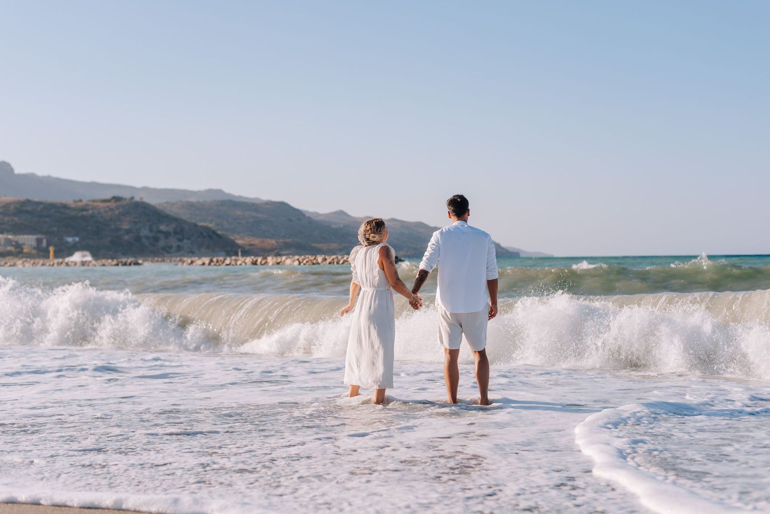 Couple in white clothes holding hands standing at ocean shoreline with mountain backdrop.