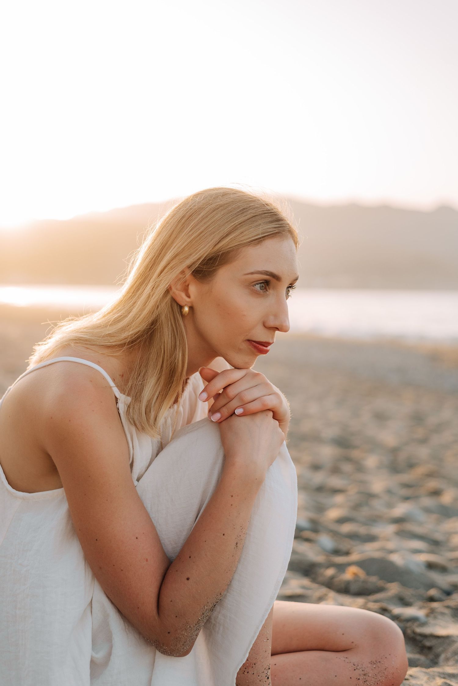 Blonde woman in white dress sitting on sandy beach gazing sideways during warm sunset light.