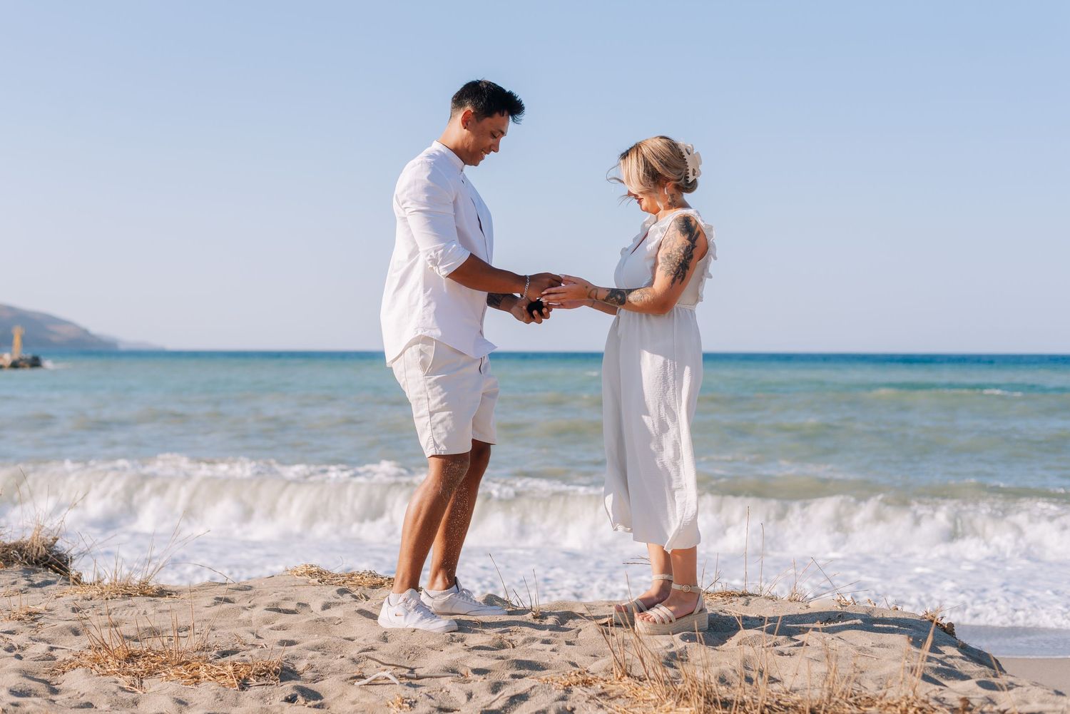 Couple in white outfits holding hands on a sunny beach with ocean waves in the background.