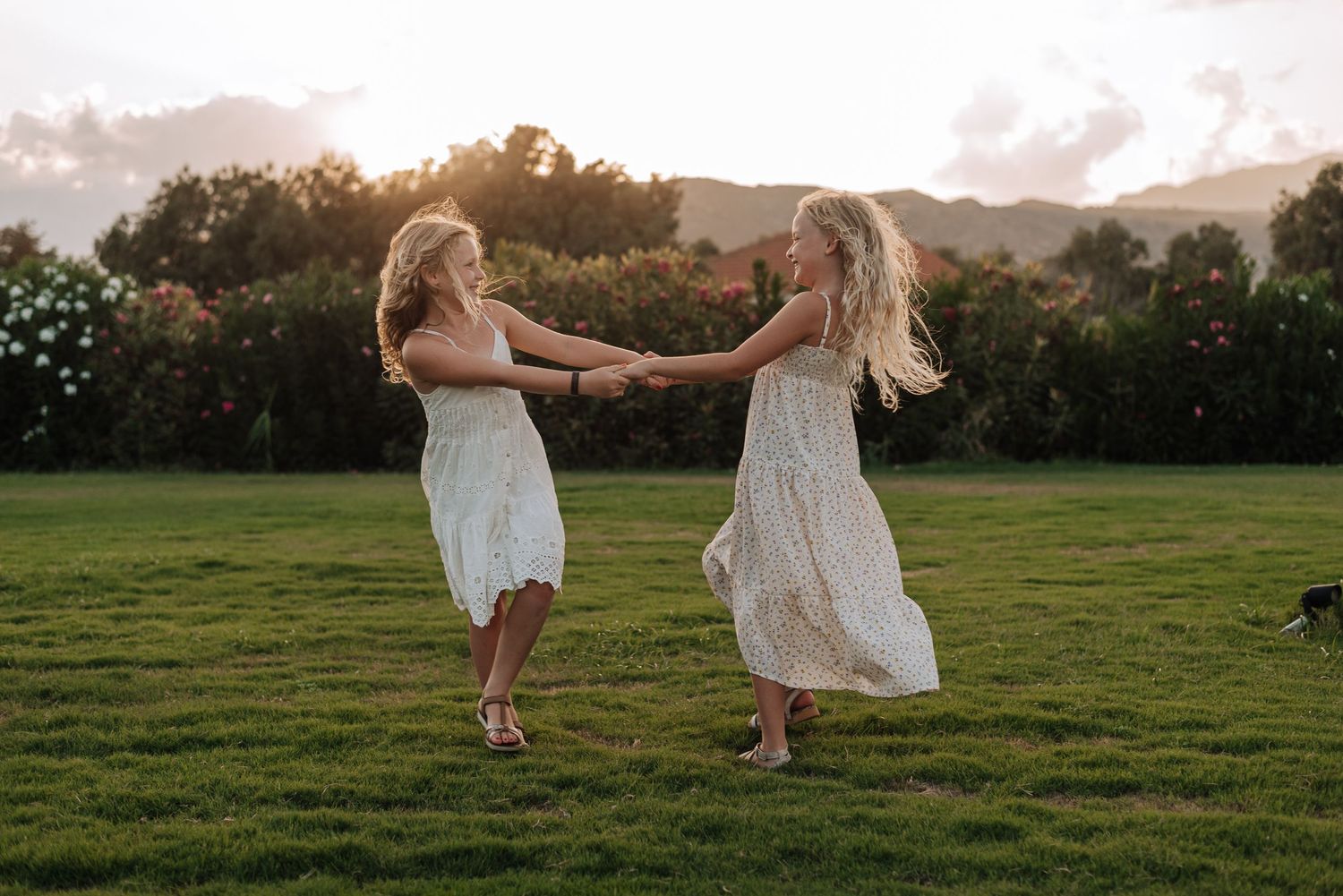 Two girls in white dresses hold hands and spin joyfully on a green lawn at golden hour with mountains in the background.