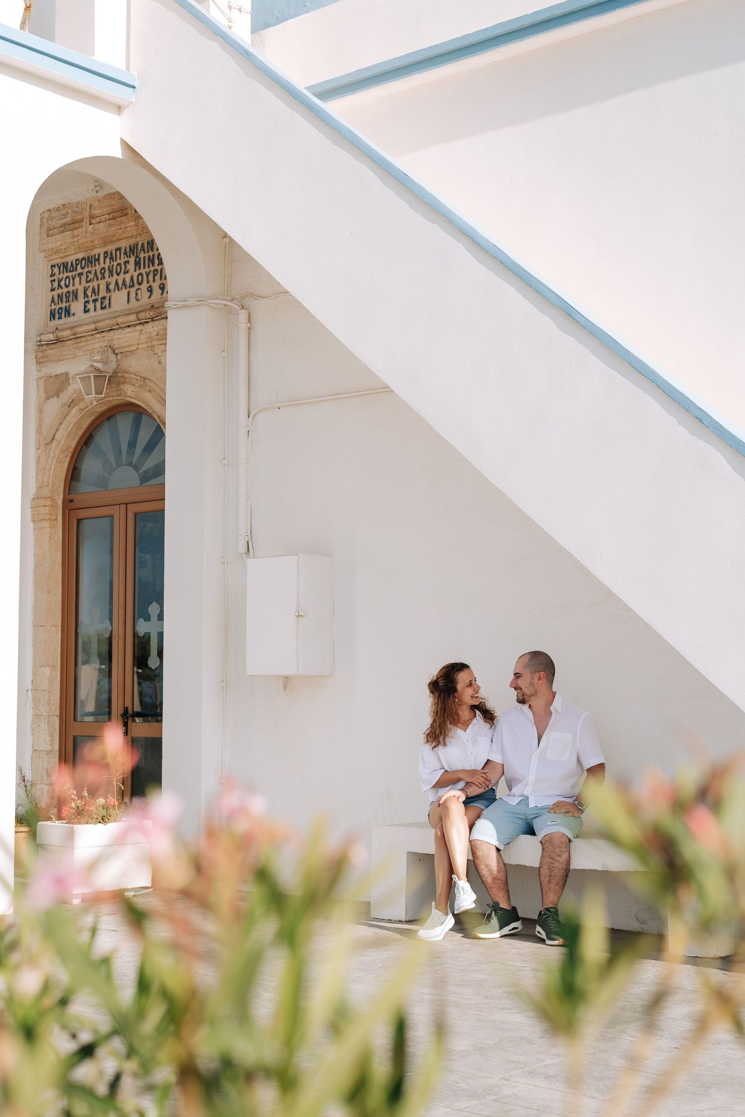 Couple sitting together under white staircase outside a Mediterranean building with arched doorway.