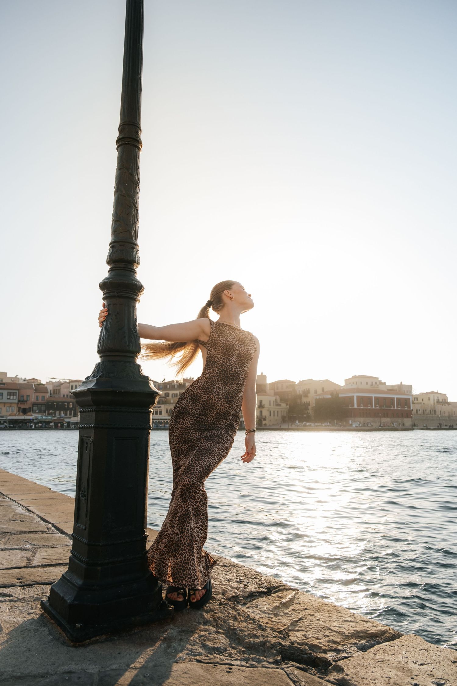 Woman in leopard print maxi dress leaning on lamppost at golden hour by waterfront.