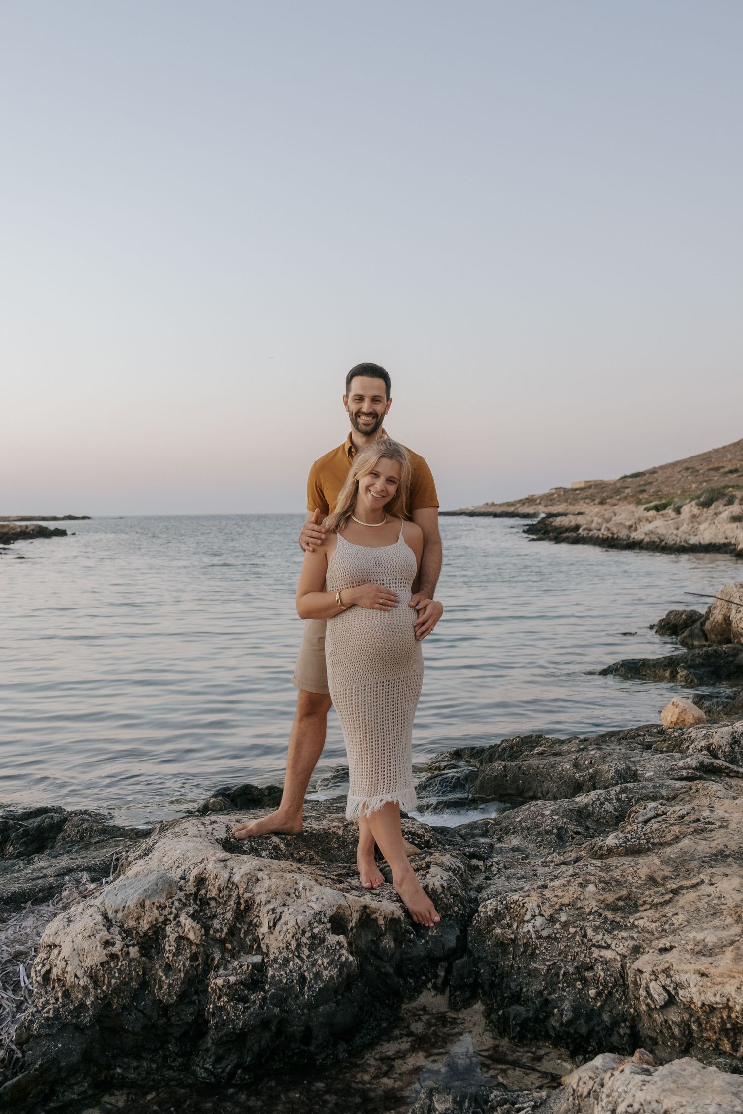 Pregnant couple posing on rocky shoreline at sunset, man in brown shirt embracing woman in cream ribbed dress.