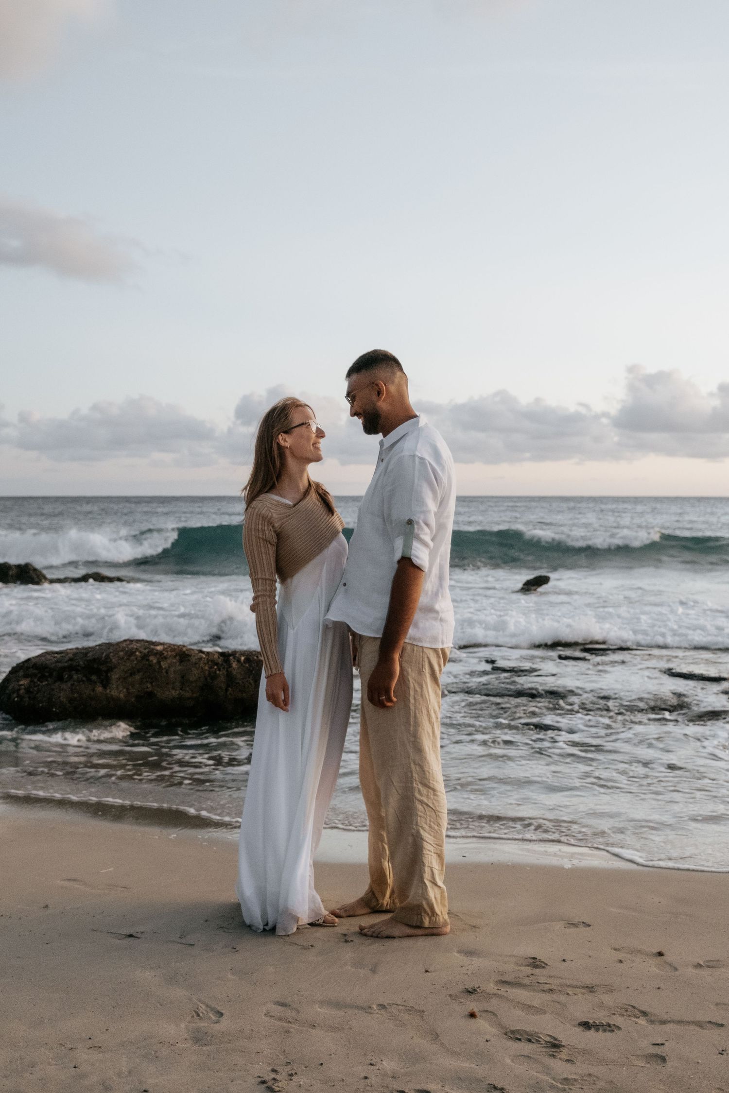 Couple standing face to face on a sandy beach at sunset with dramatic ocean waves crashing on rocks behind them.