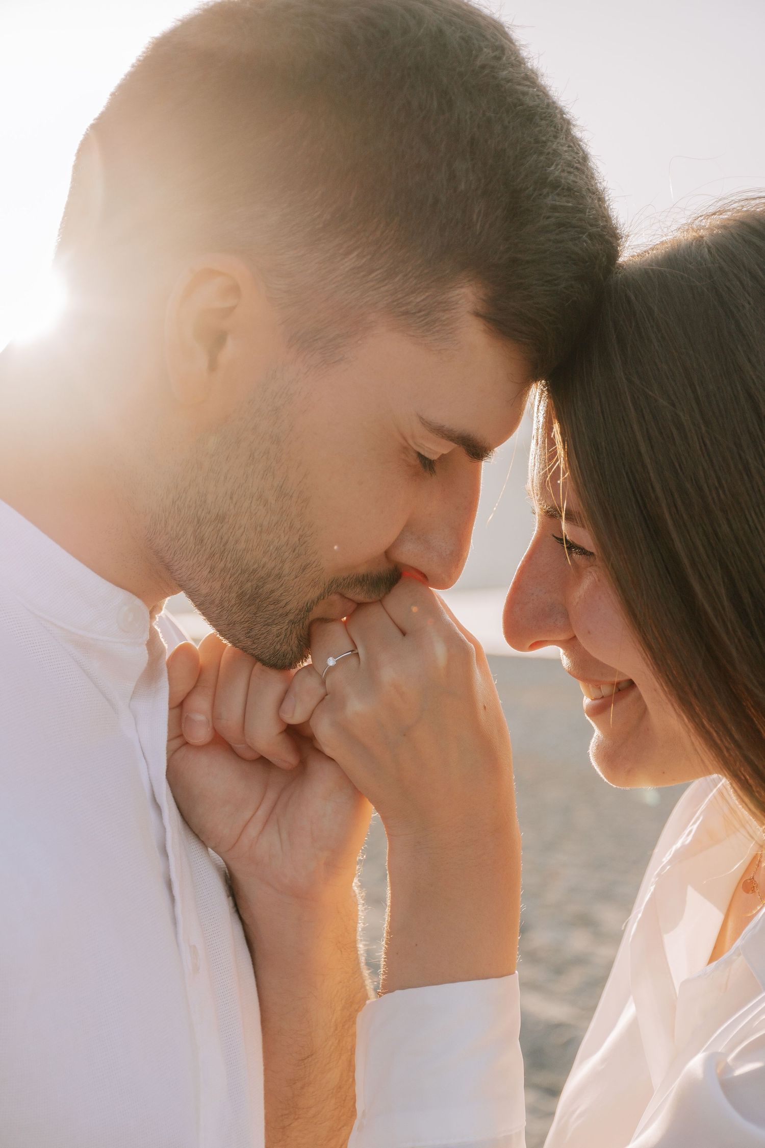 Couple sharing an intimate moment in golden backlight, man kissing woman's hand near the beach.
