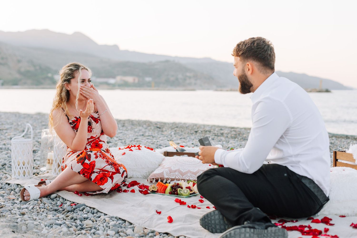 Man proposing to surprised woman on a beach blanket surrounded by red rose petals at sunset.