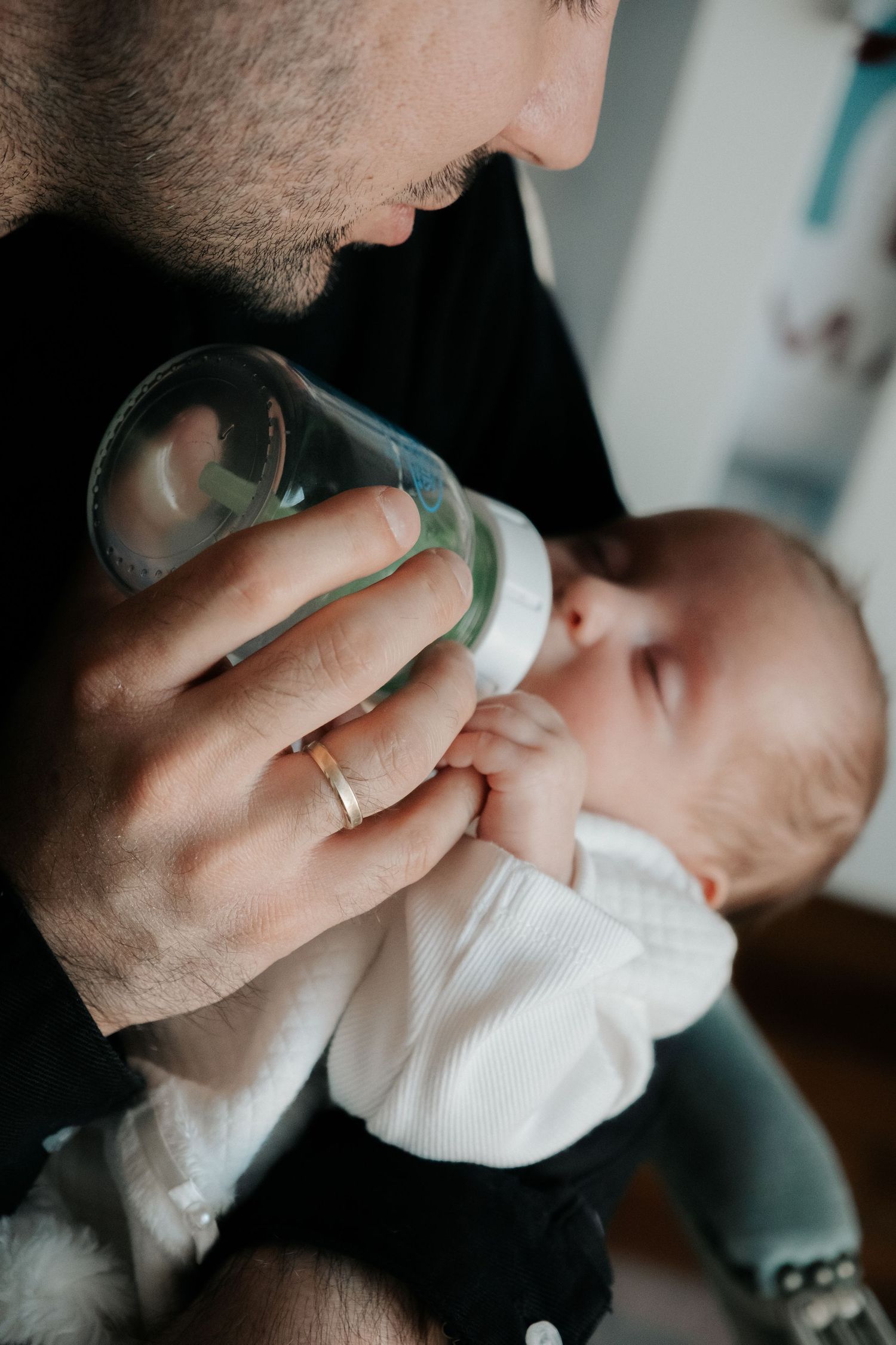 Father with wedding ring bottle-feeding his newborn baby while looking down lovingly at the infant.