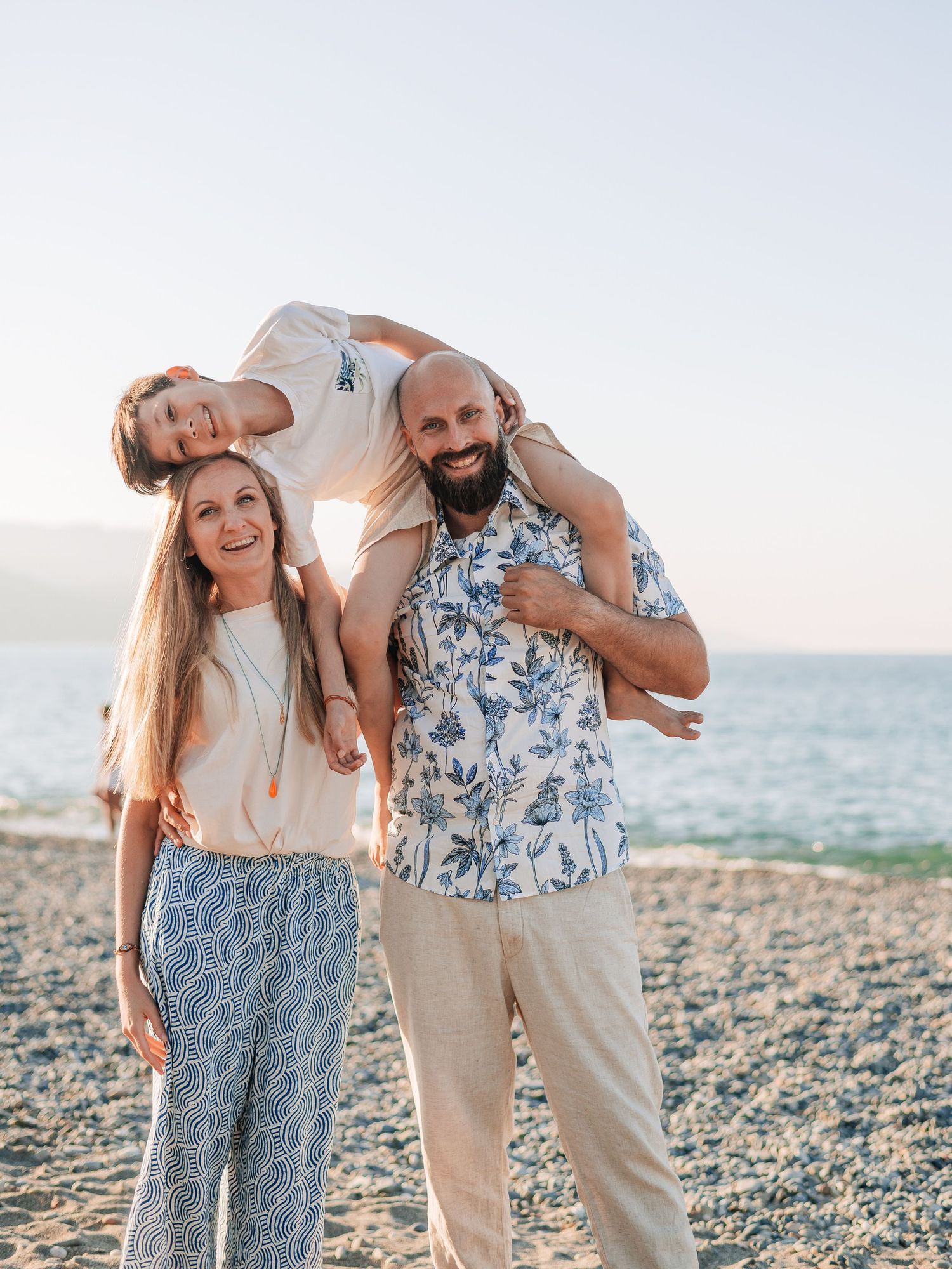 Happy family of three posing on a rocky beach at sunset, father carrying child on shoulders.