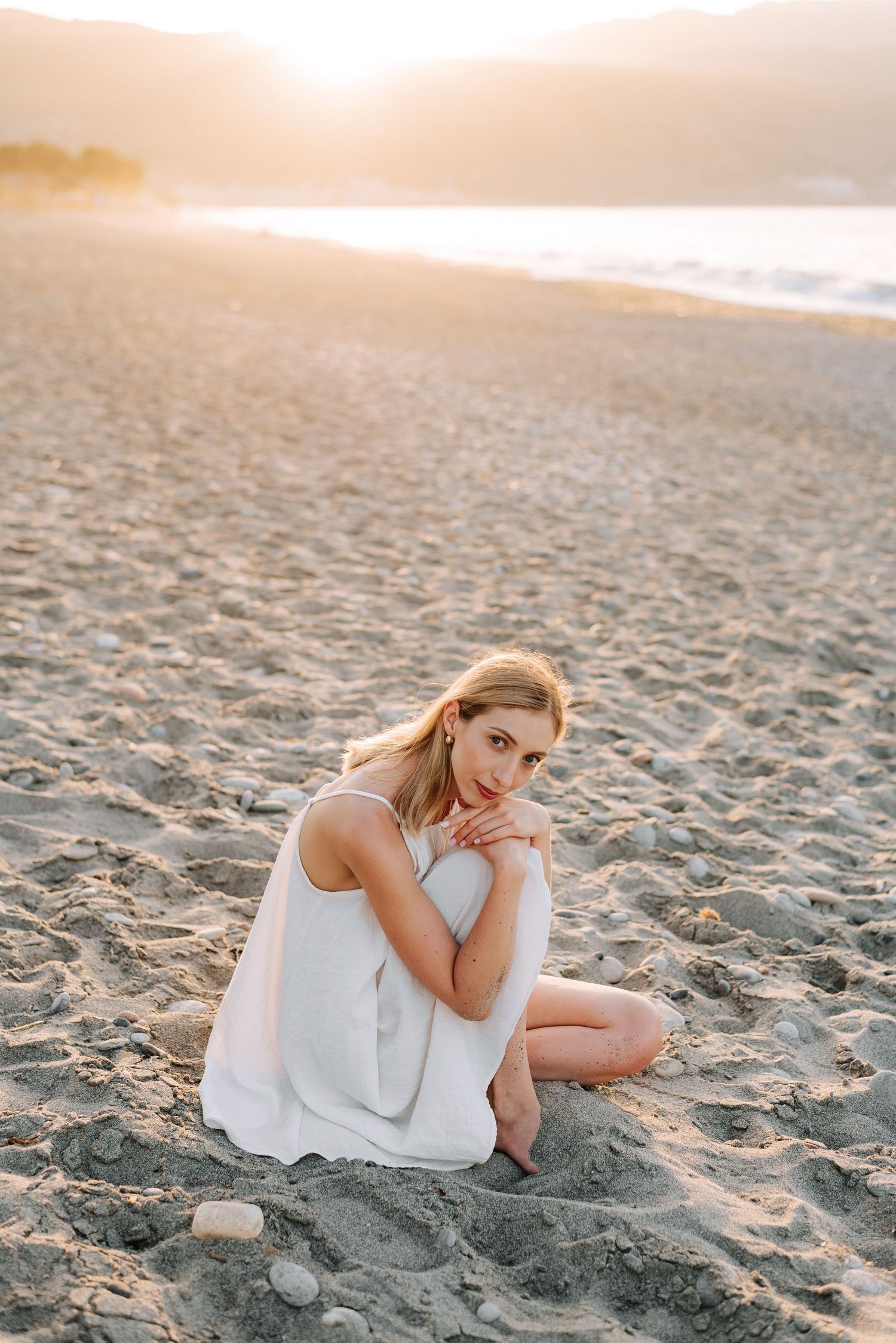 Blonde woman in white dress sitting on sandy beach at golden sunset by the ocean.