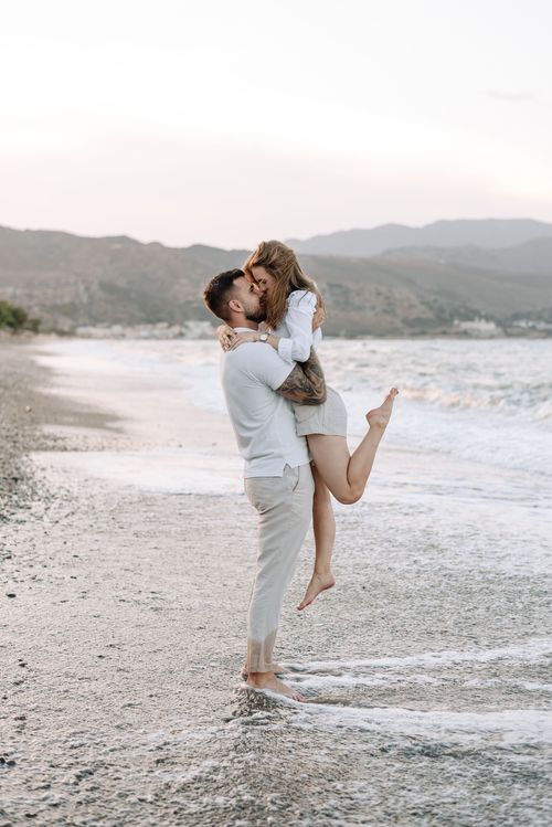 Romantic couple embracing on a pebble beach at sunset with mountains in the background.