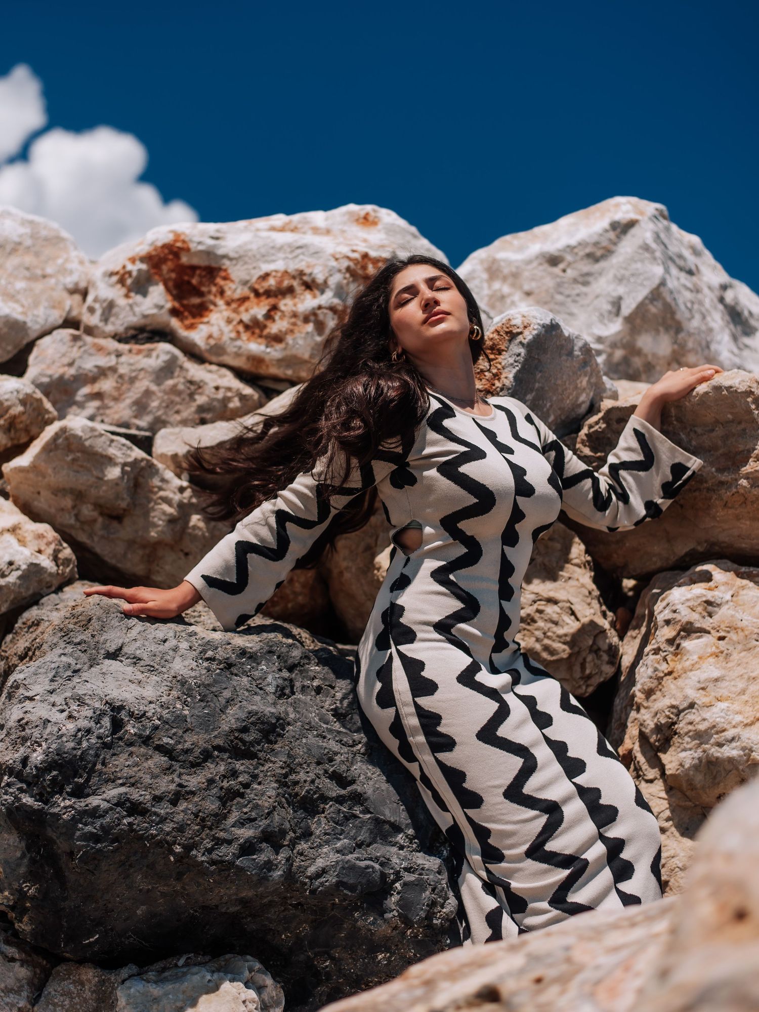 Woman in black and white geometric print dress leaning back on sun-drenched rocky boulders.