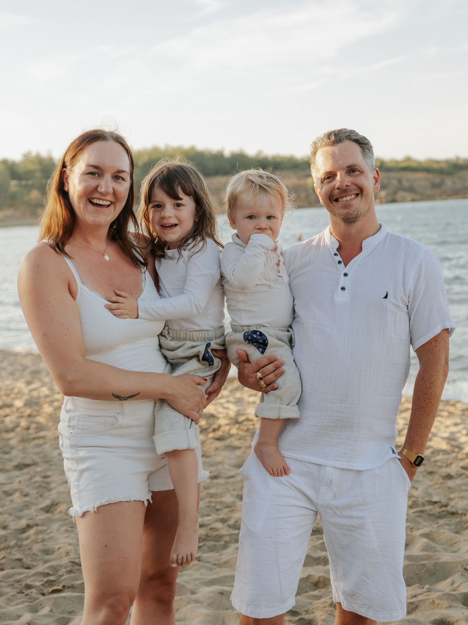 Happy family of four posing on sandy beach in white outfits at golden hour.
