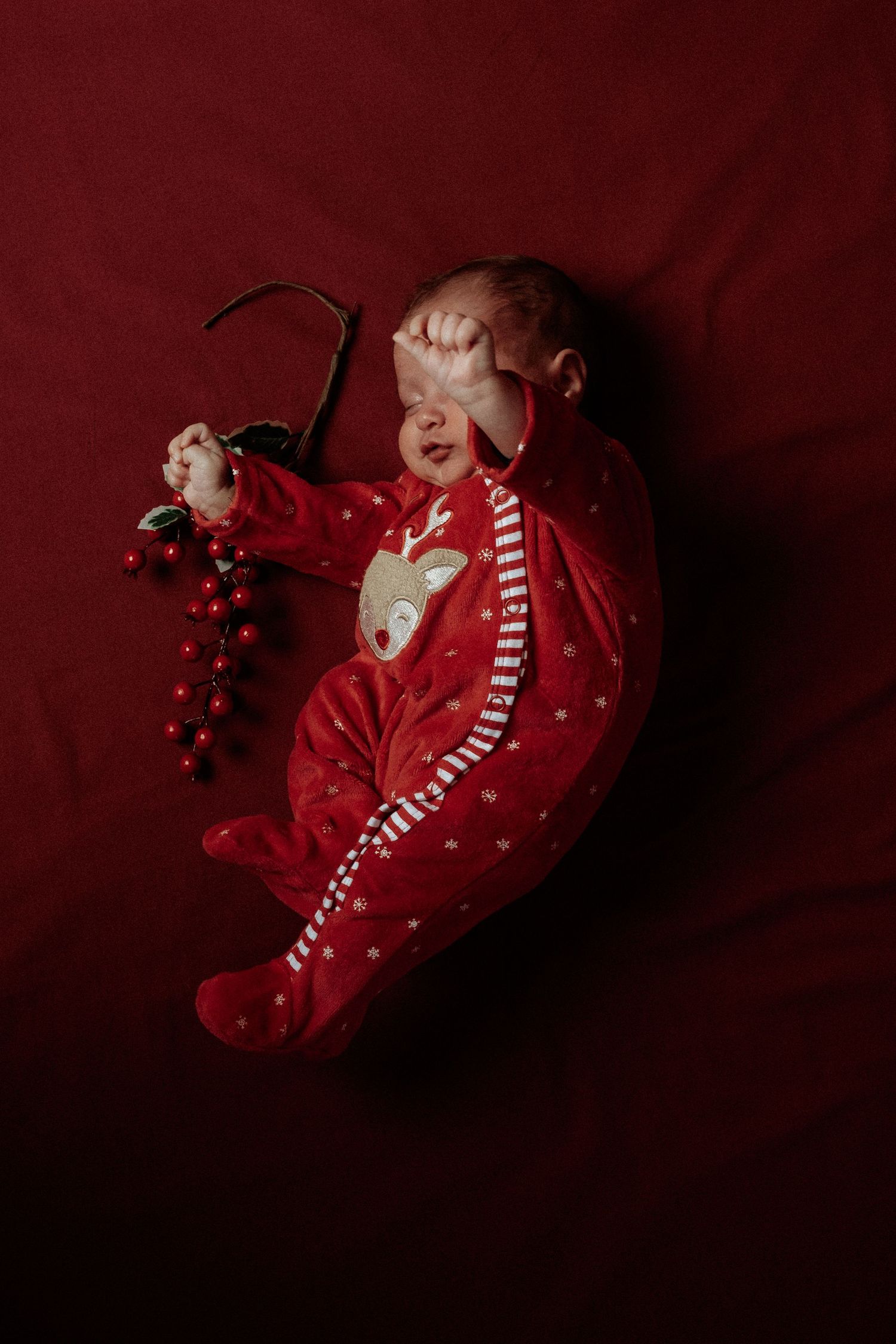 Newborn baby in red reindeer Christmas pajamas lying on red fabric holding a festive holly decoration.