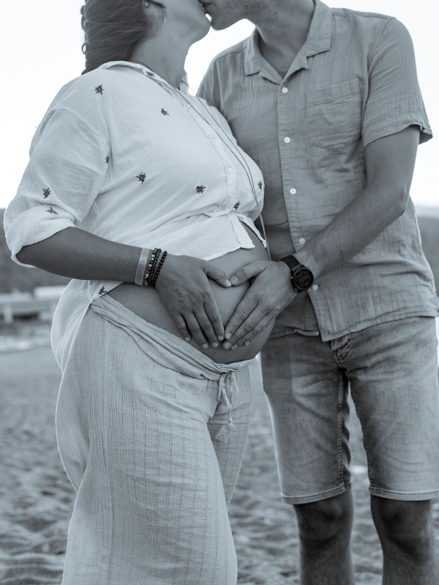 Black and white photo of couple making heart shape on pregnant belly at beach.