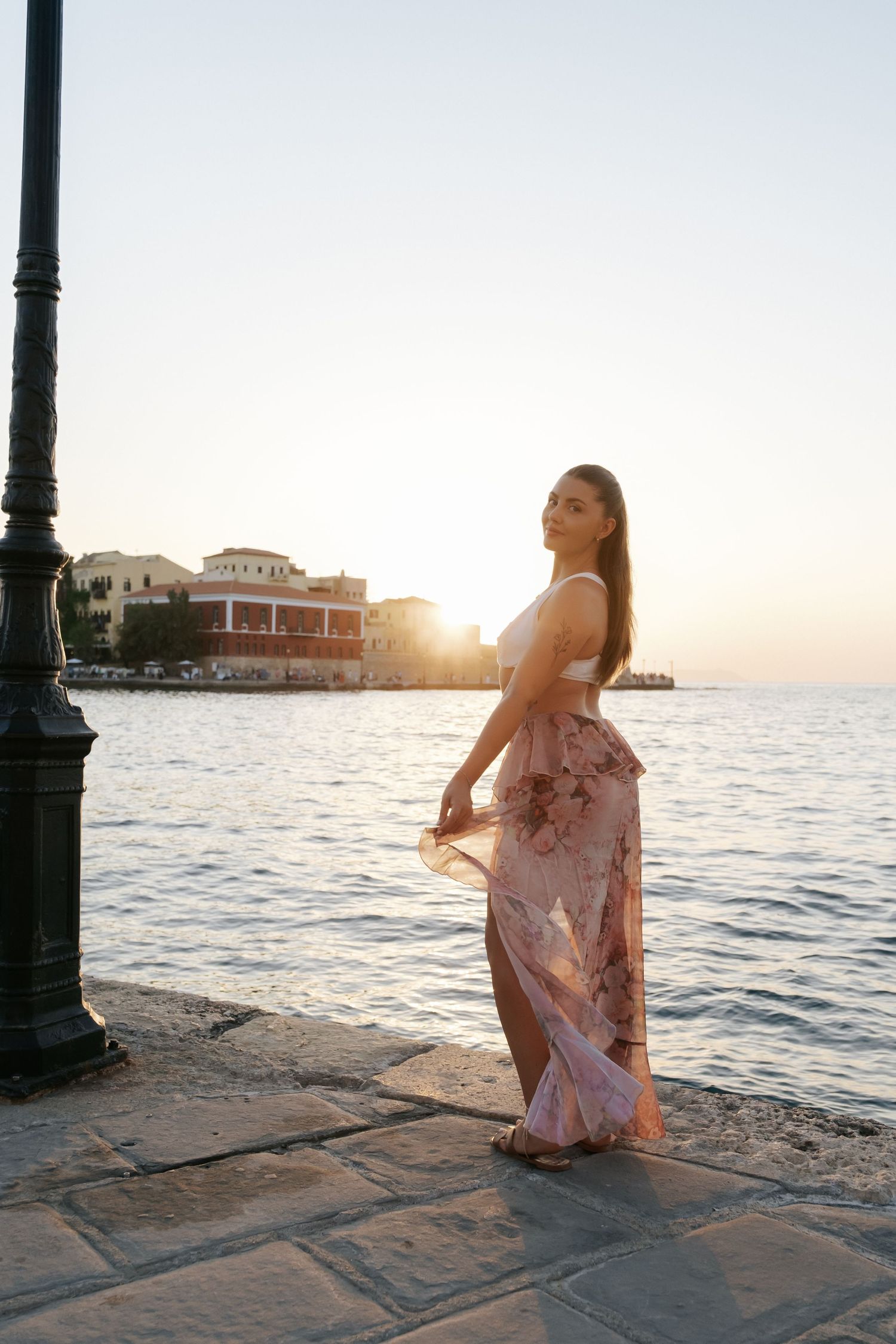 Woman in floral skirt and white top standing by seaside lamppost at golden sunset.