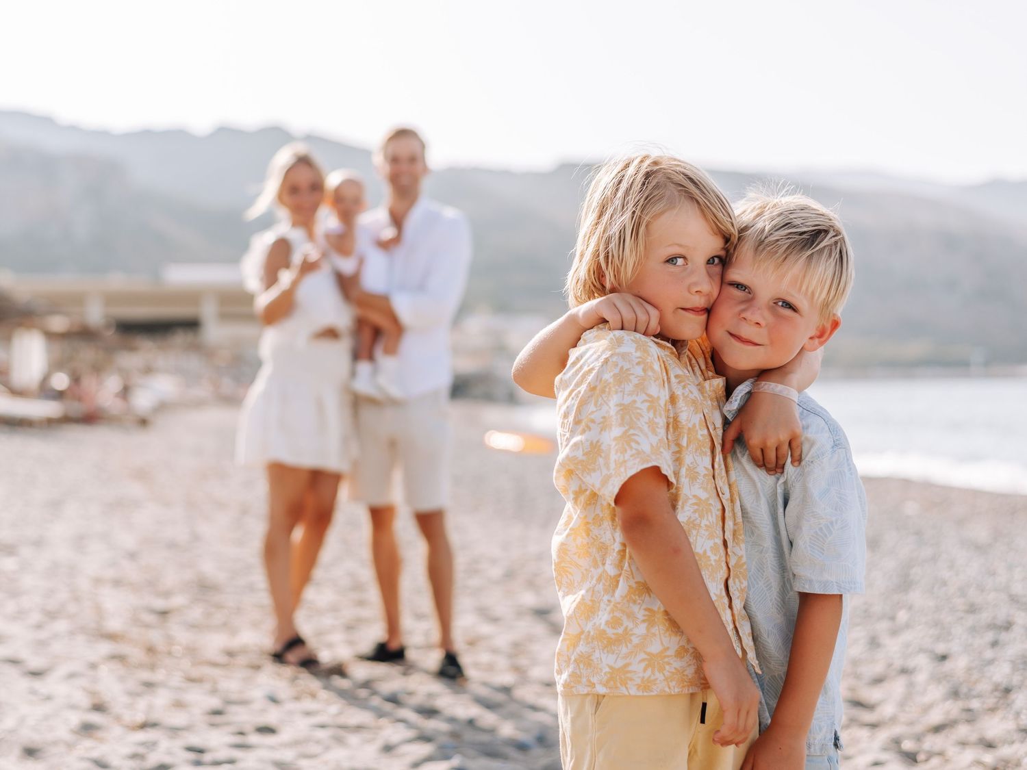Two young brothers hugging on a beach with their parents and baby sibling standing behind them.