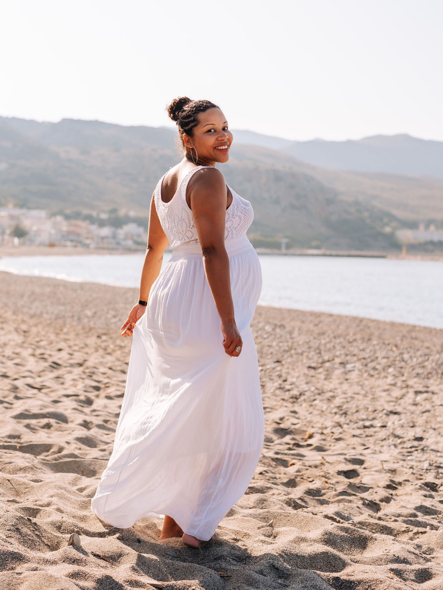 Pregnant woman in flowing white maxi dress walking barefoot on sandy beach with mountains in background.