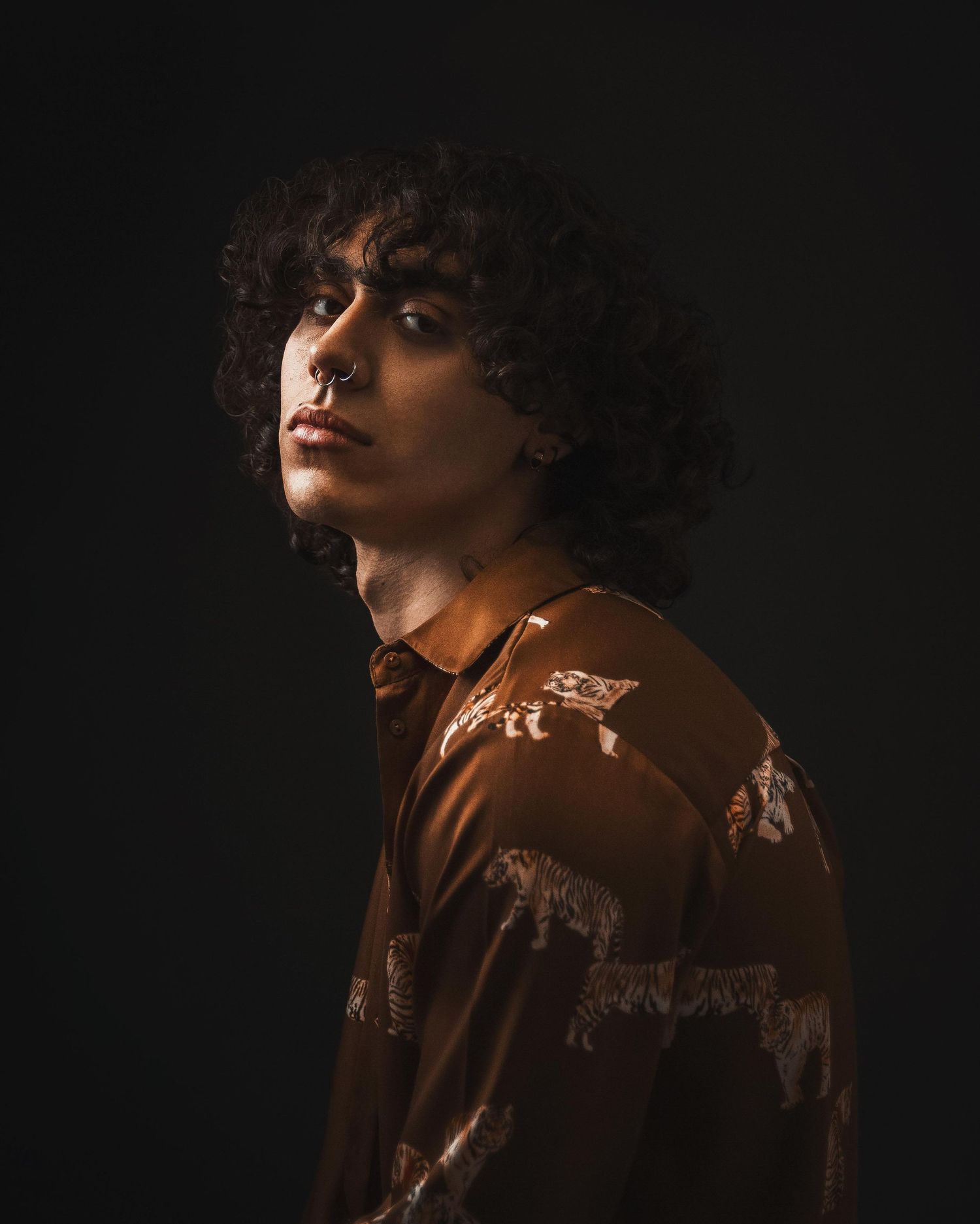 Young man with curly hair and nose ring wearing brown patterned shirt against dark background.