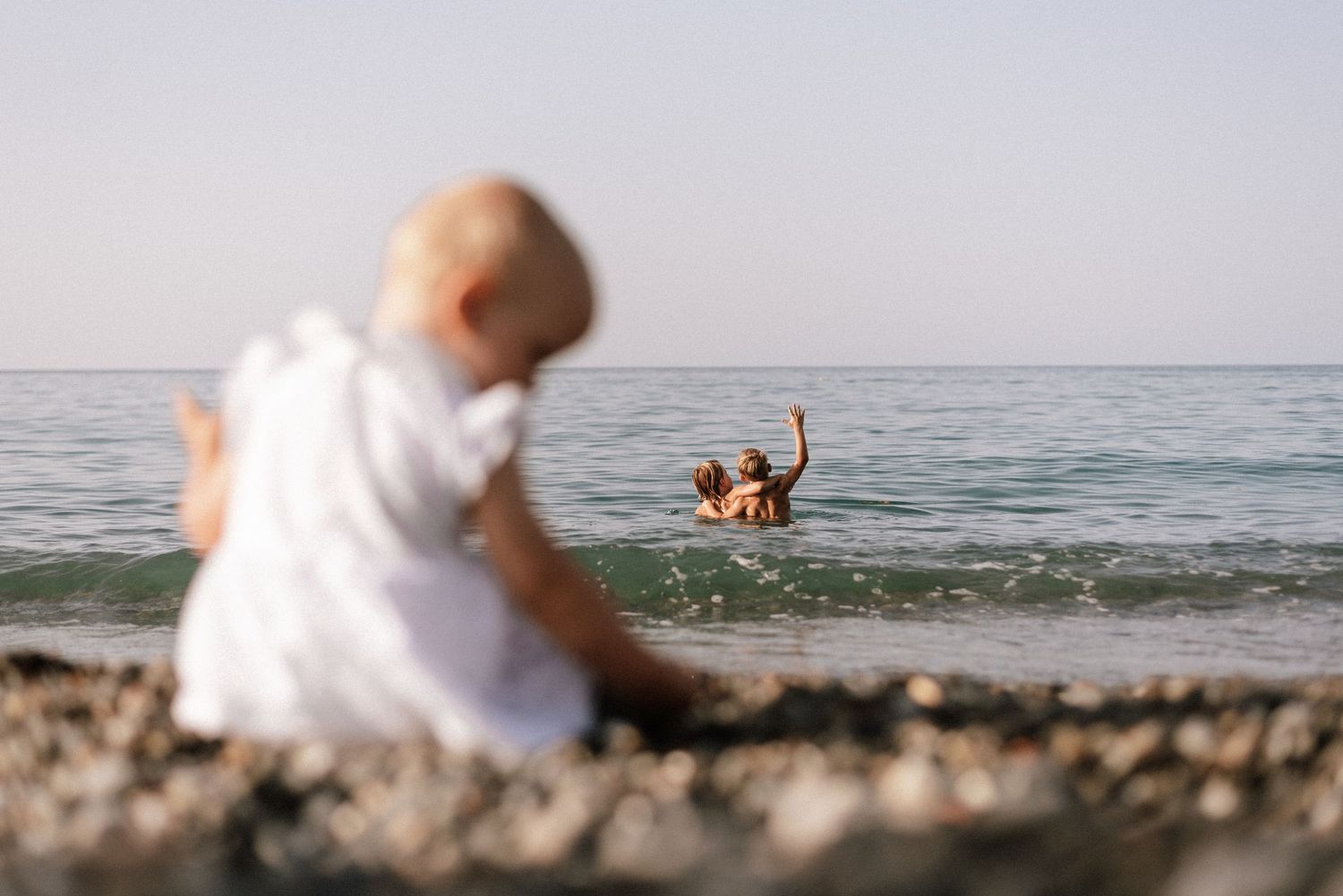 Baby in white dress sitting on rocky beach shore watching two adults playing in the ocean waves.