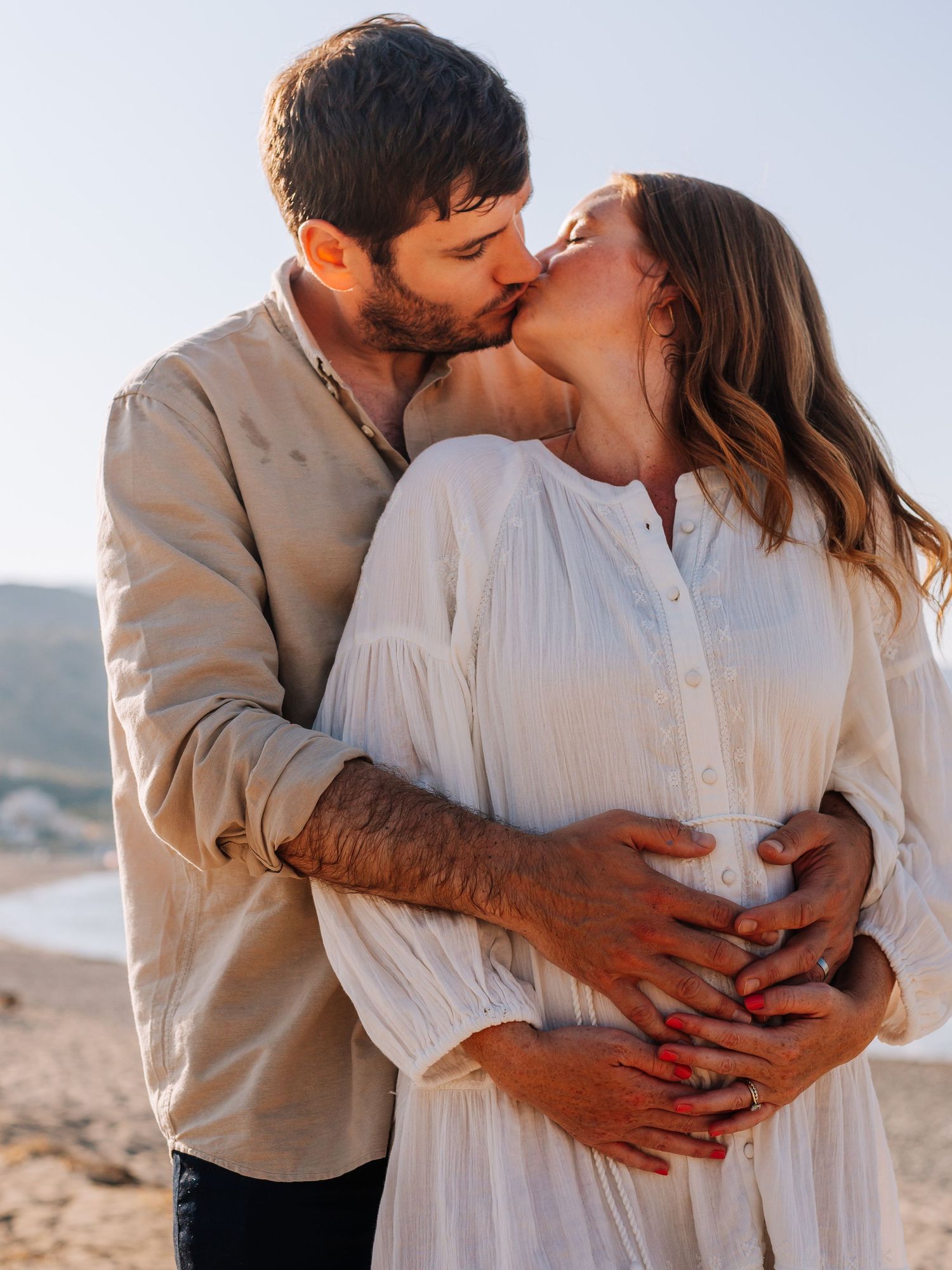A couple shares a tender kiss on a beach, with the man cradling the pregnant woman's belly in a romantic maternity photo.