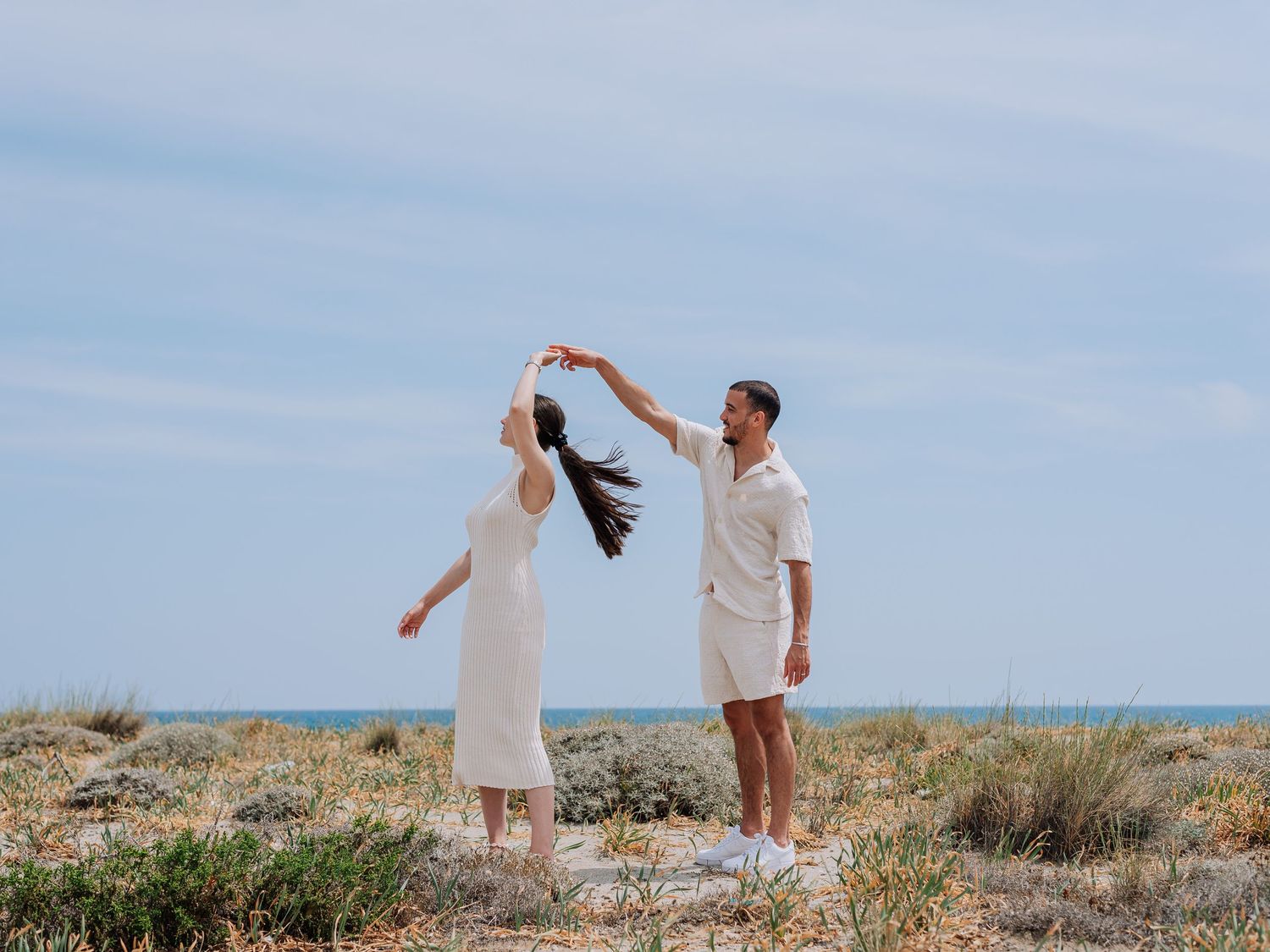Couple dancing together on a coastal shrubland with the blue ocean and cloudy sky in the background.