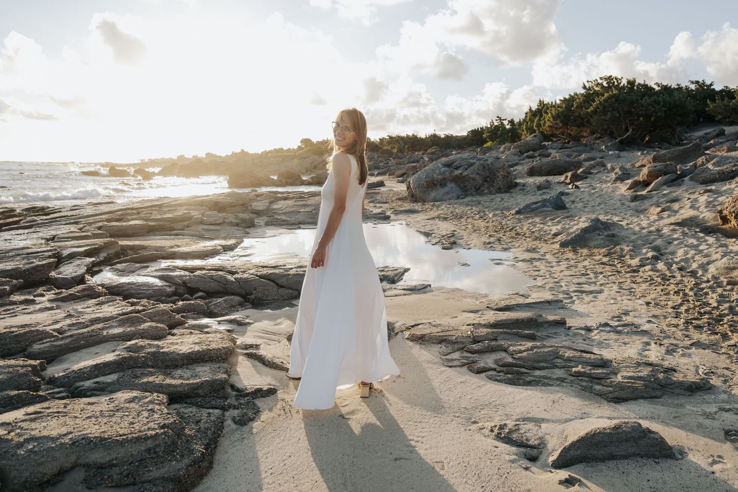 Bride in white flowing gown walking on sandy rocky beach during golden sunset hour.