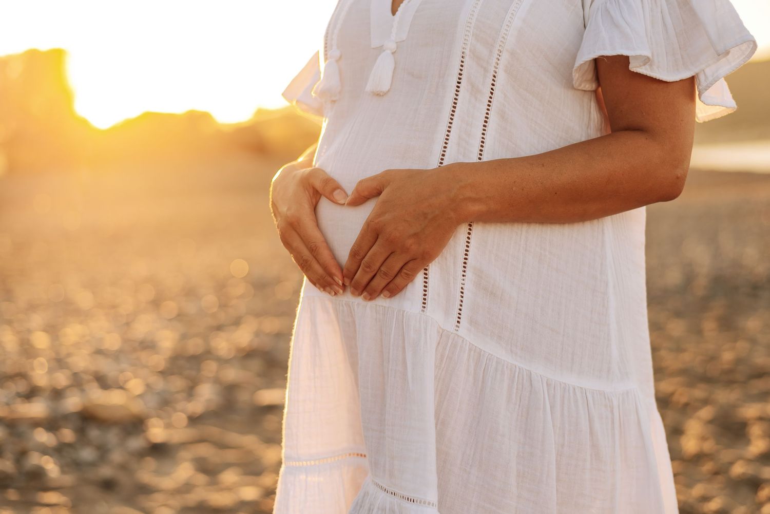 Pregnant woman in white dress forming heart shape on baby bump at golden sunset on beach.