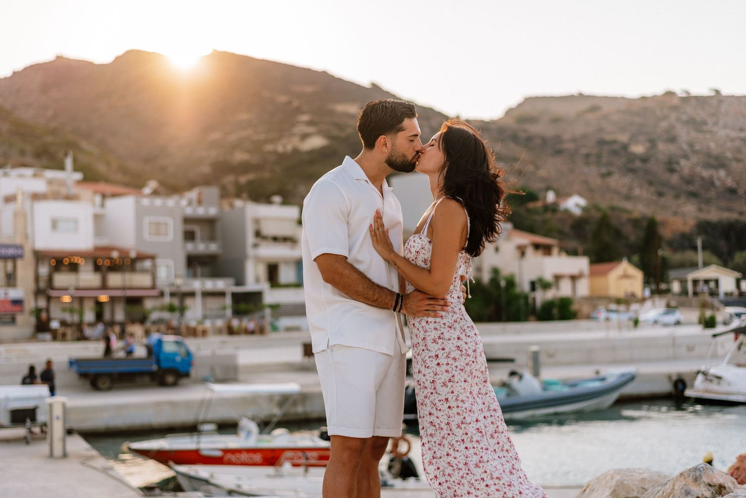 Couple sharing a kiss at a Mediterranean harbor with boats and mountains at golden sunset.