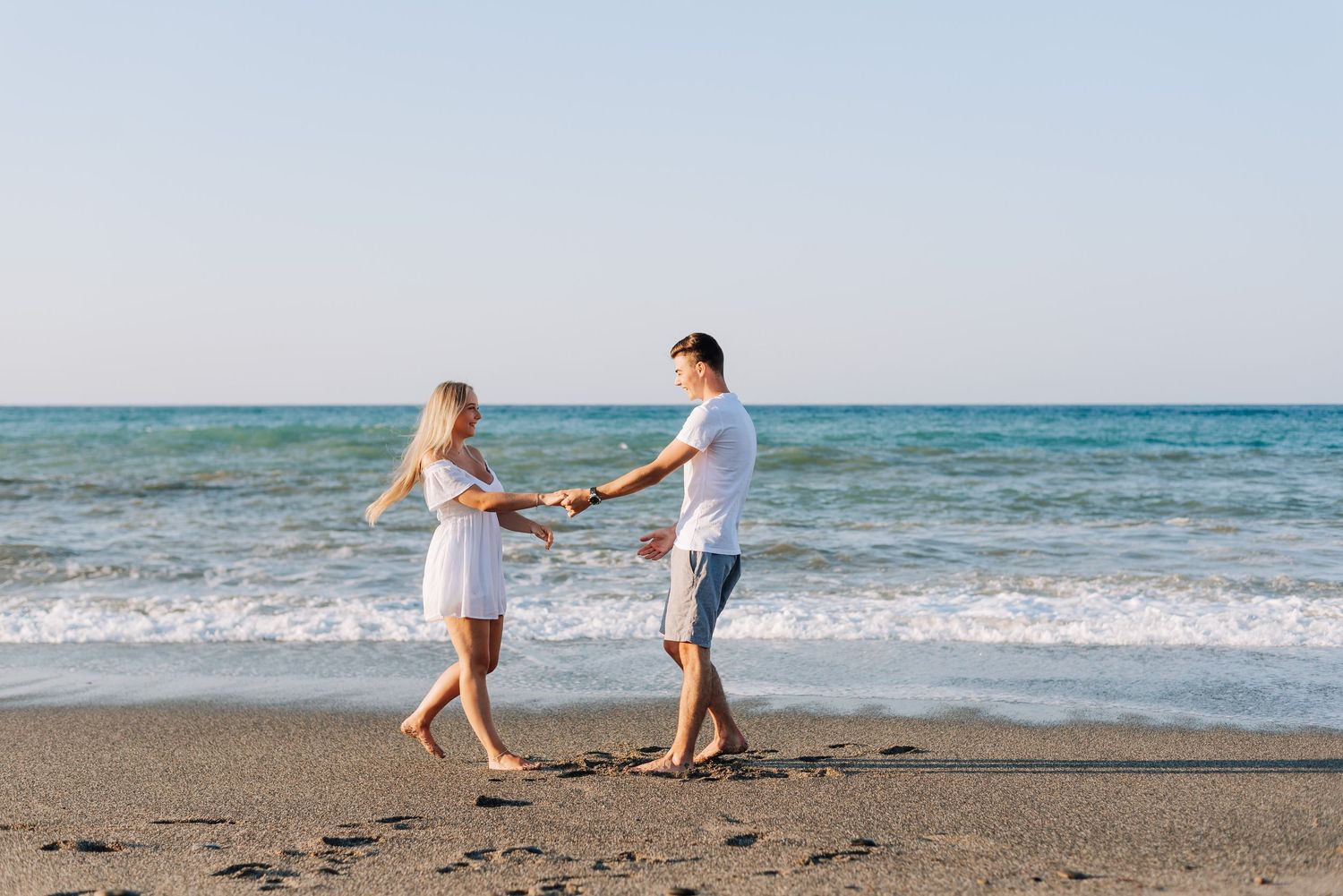 Couple holding hands and dancing on a sunny beach with turquoise ocean waves in the background.