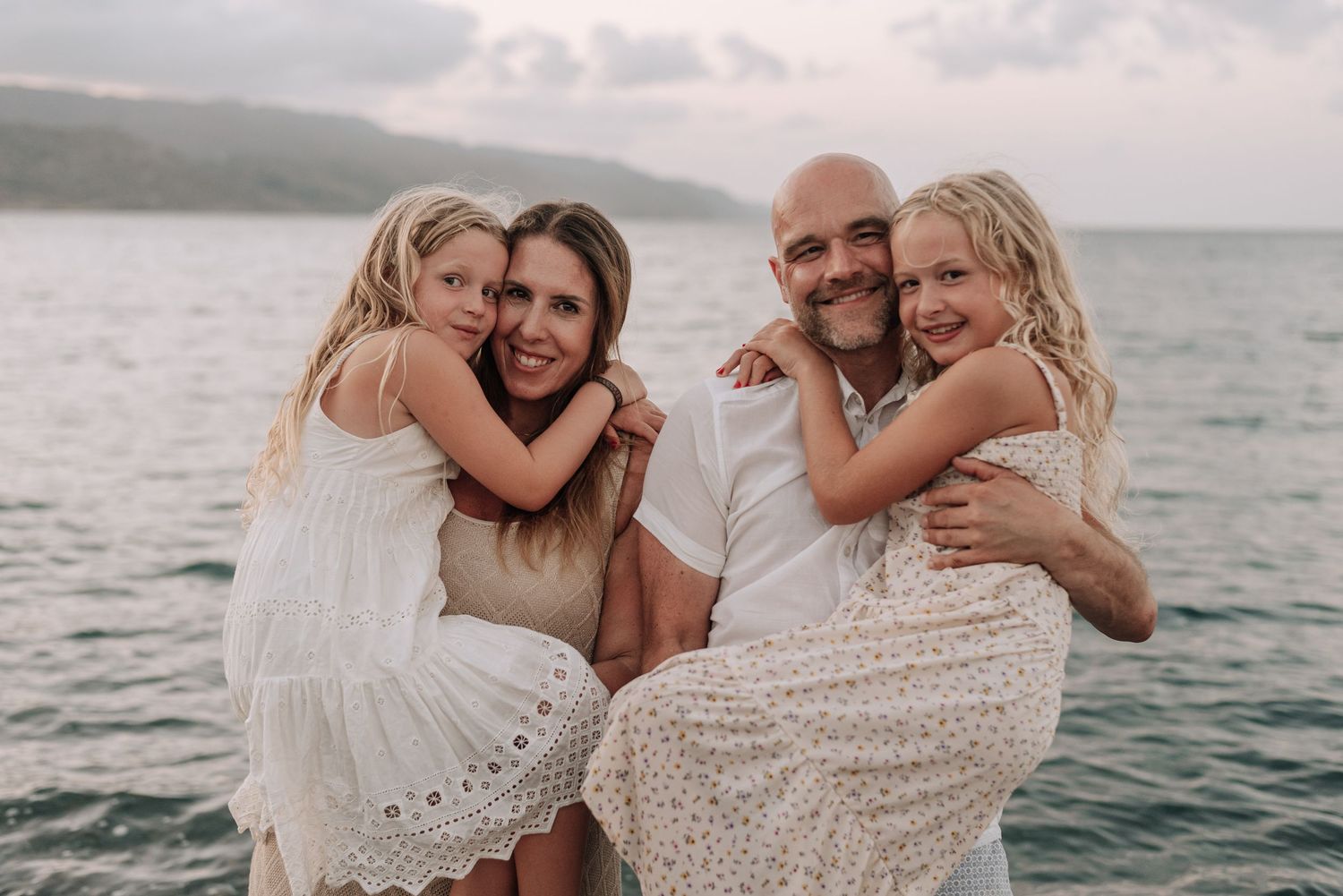 Happy family of four embracing near the ocean at dusk, parents holding daughters.
