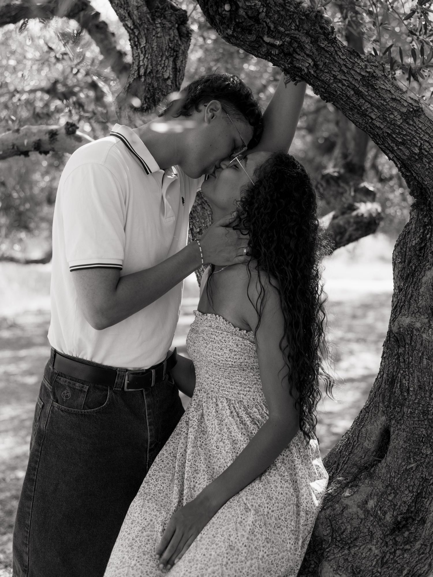 Black and white romantic photo of a couple kissing under a twisted tree in a garden setting.