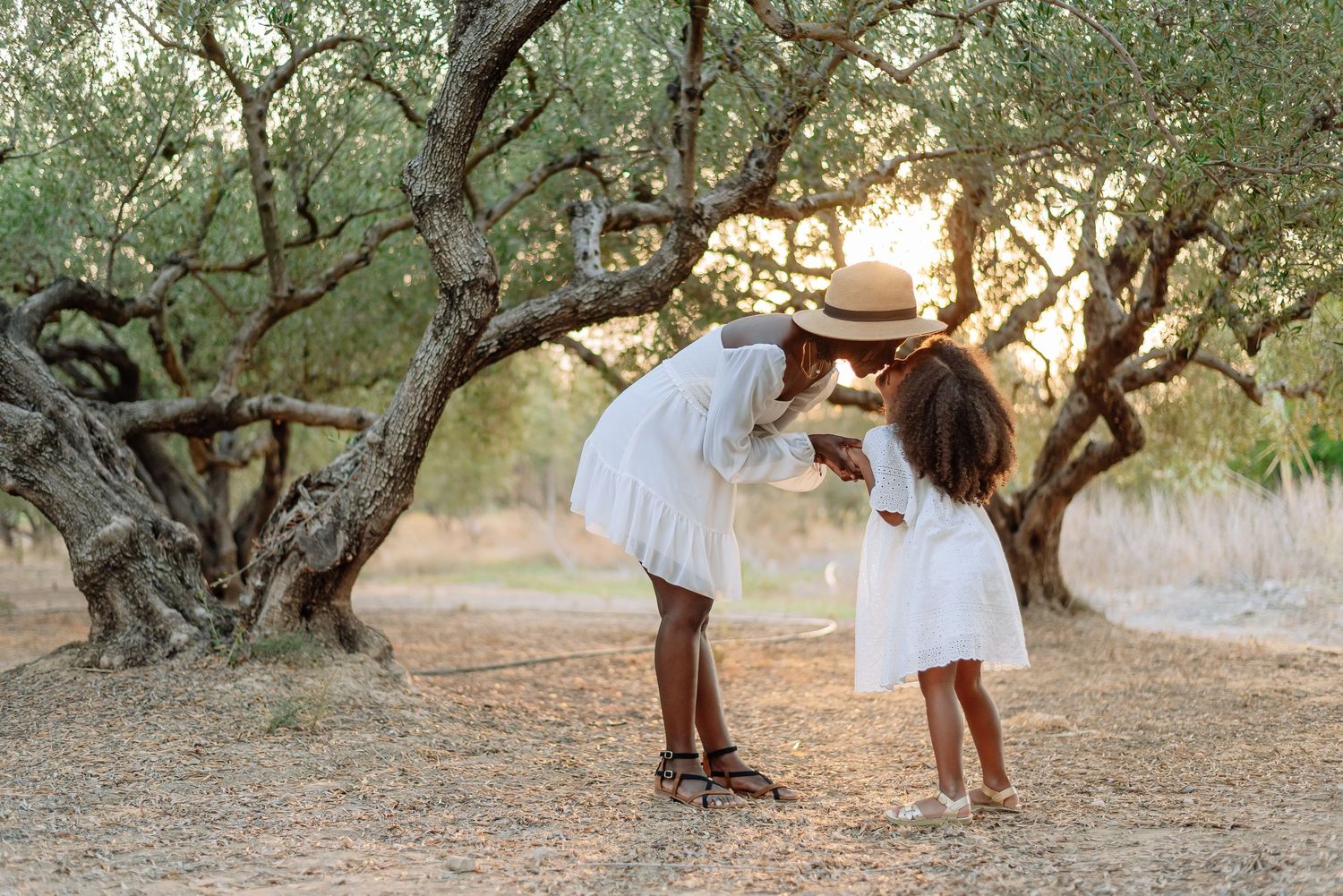 Two children in white dresses share a tender moment under olive trees at golden hour.