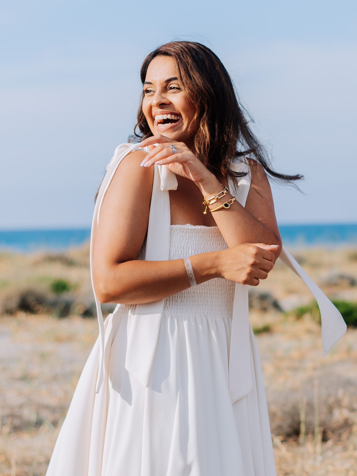 Joyful woman in white smocked sundress with tie shoulders laughing on sunny coastal beach.