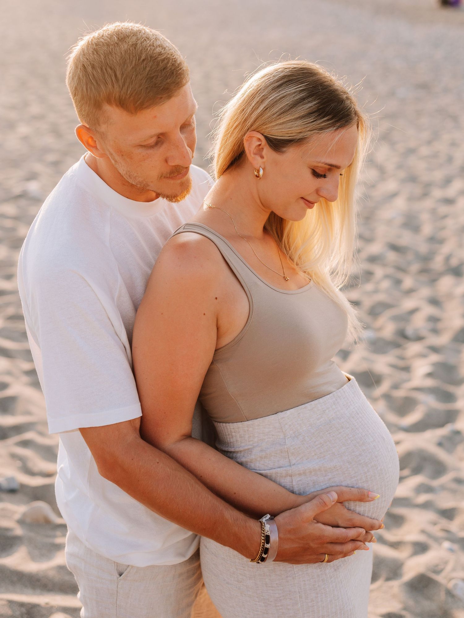 Couple on sandy beach at sunset, man embracing pregnant woman from behind, both looking down at belly.