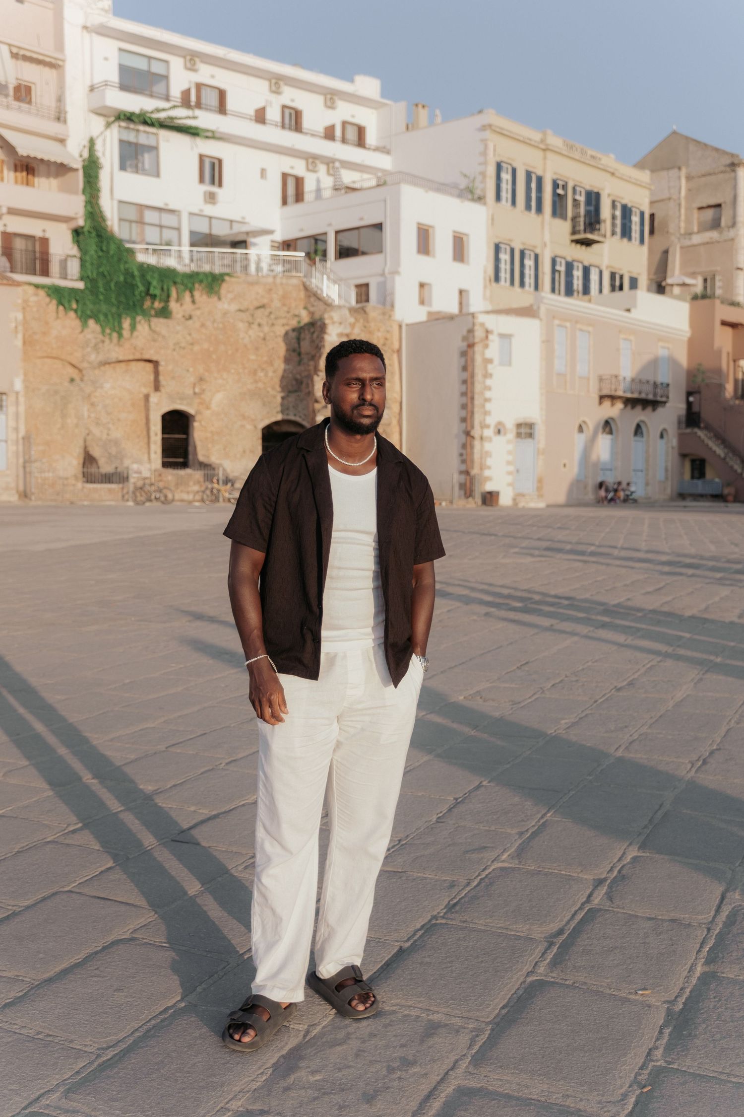 Confident Black man in white pants and dark shirt posing in sun-drenched European coastal square.